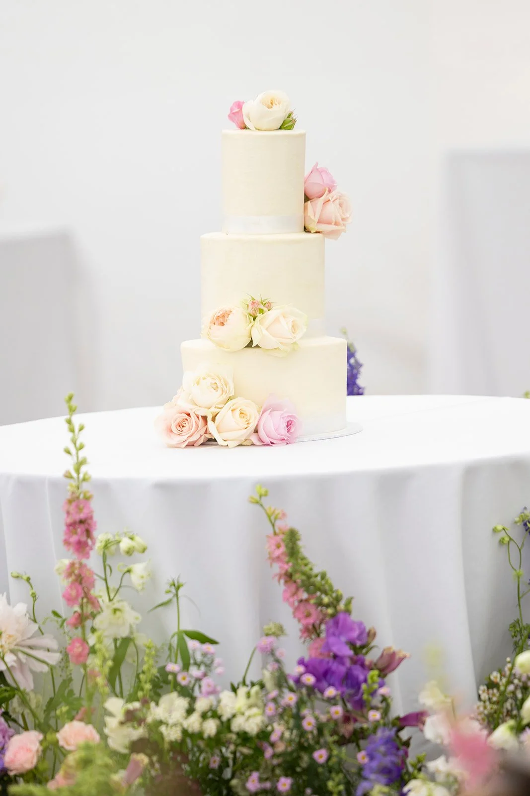 Three-tiered white wedding cake decorated with pastel pink, white, and cream roses, placed on a table with floral arrangements in the foreground.