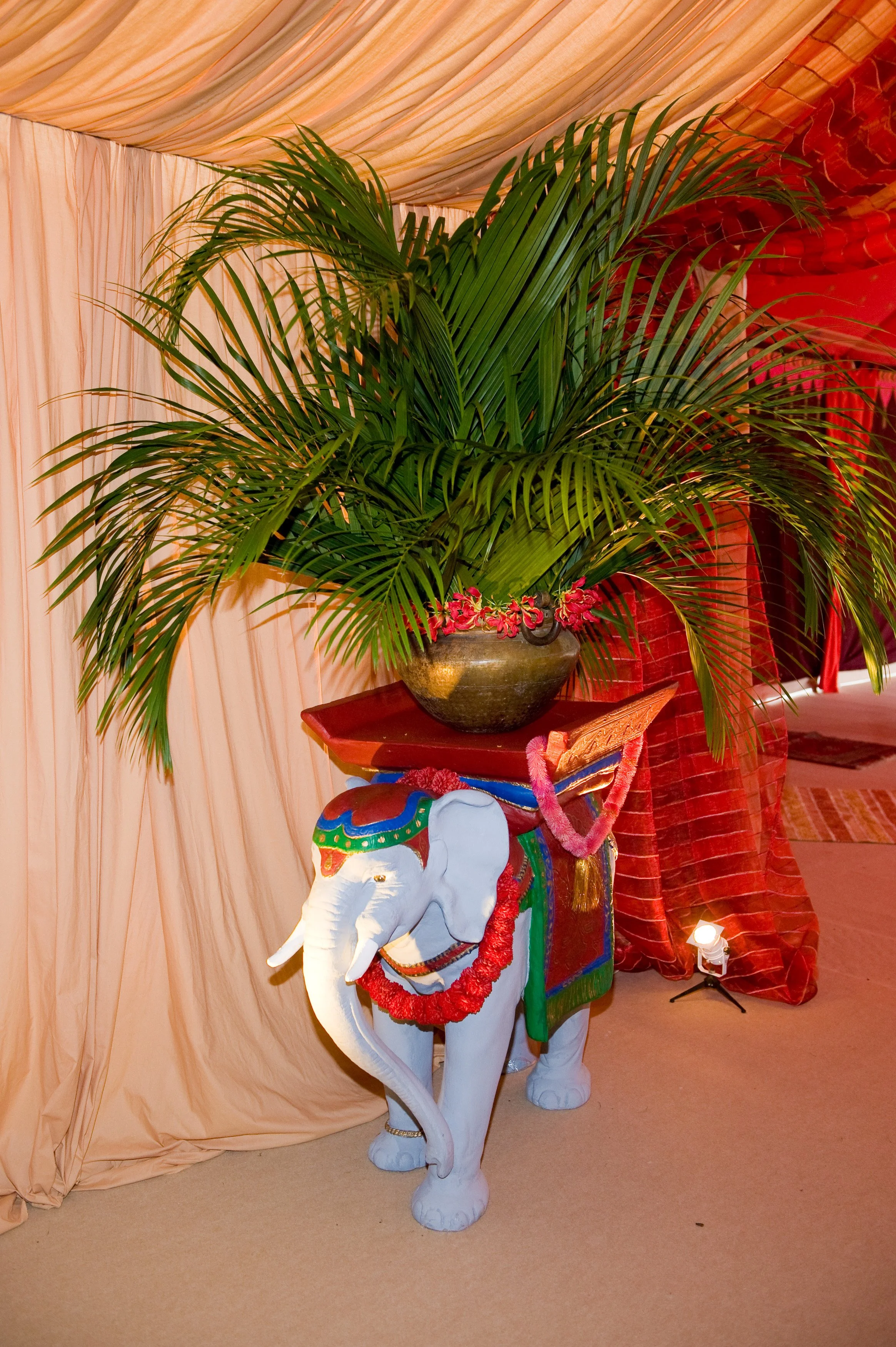 Decorative display featuring a white elephant statue with colored accents and a leafy plant in a pot on top of it, against a backdrop of tan and red curtains illuminated with soft lighting.