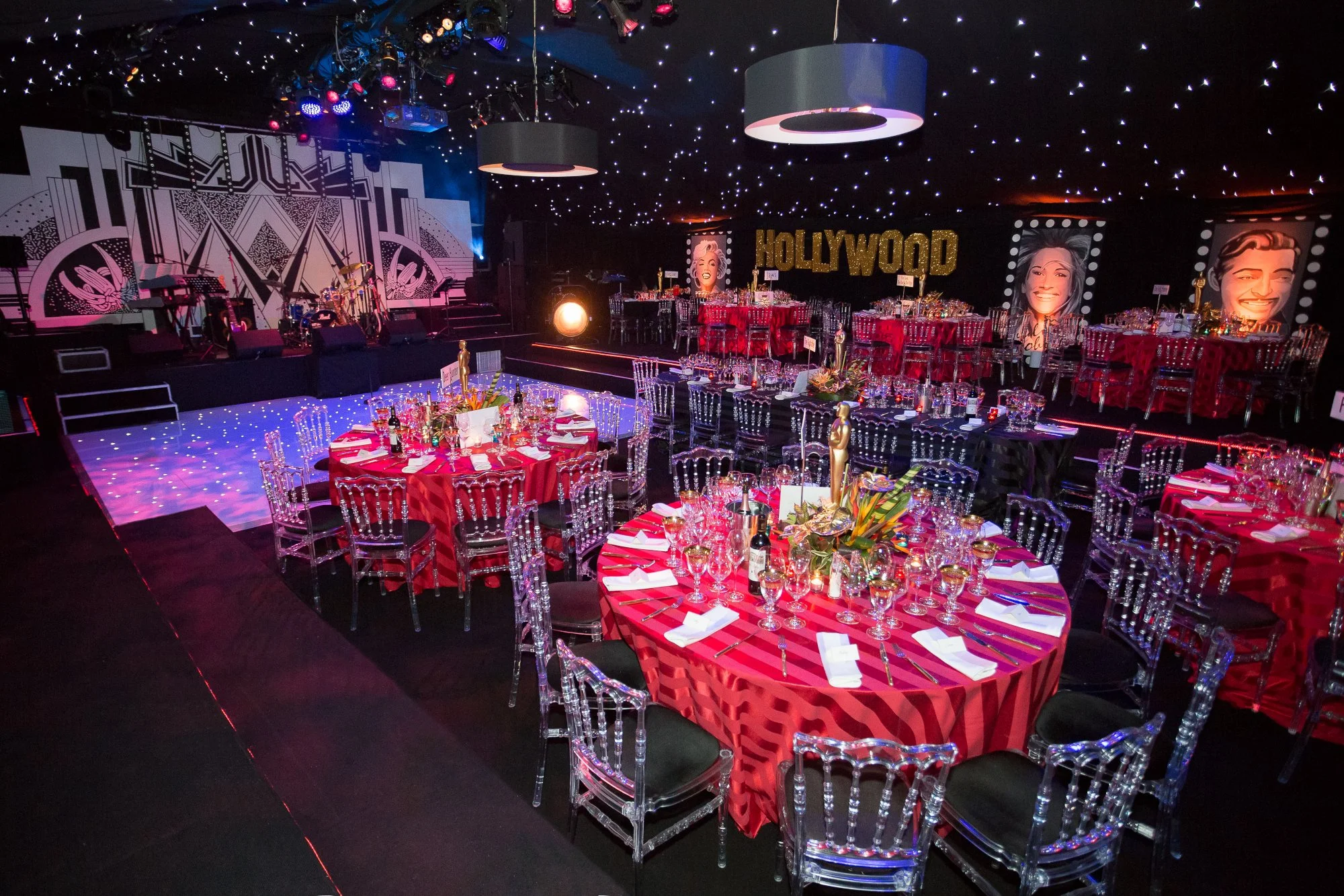 A decorated event space with round tables covered in red tablecloths, set with glassware, plates, and utensils. Clear ghost chairs surround the tables, and gold Oscar statues as centerpieces are on each table. The background features a stage with musical instruments, a large 'Hollywood' sign, and giant portrait images of smiling celebrities. The ceiling is adorned with star-like string lights and large round hanging lamps.