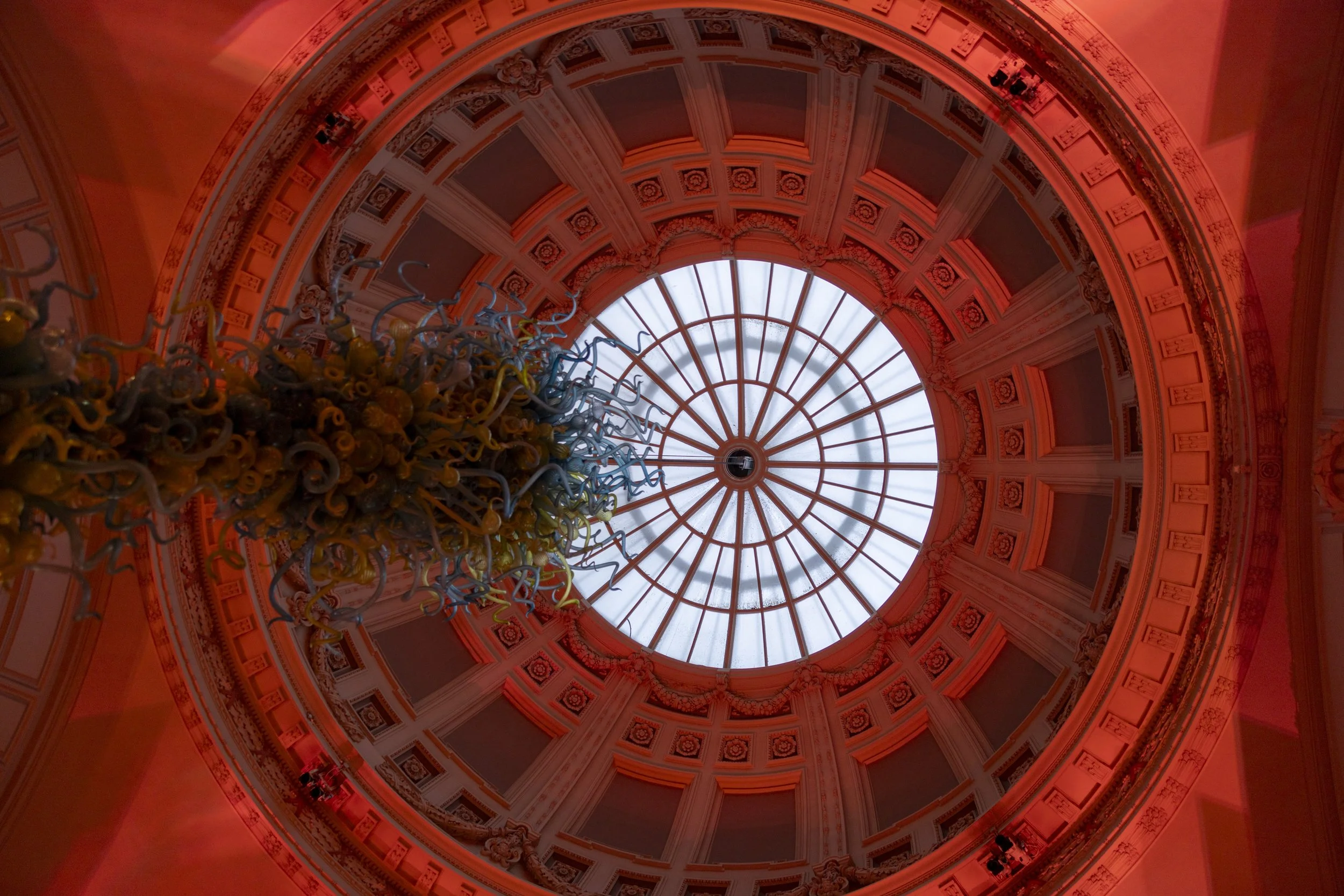 Interior view of a grand circular ceiling with intricate moldings, a large glass dome, and a chandelier at the center. The lighting gives the space a reddish hue.