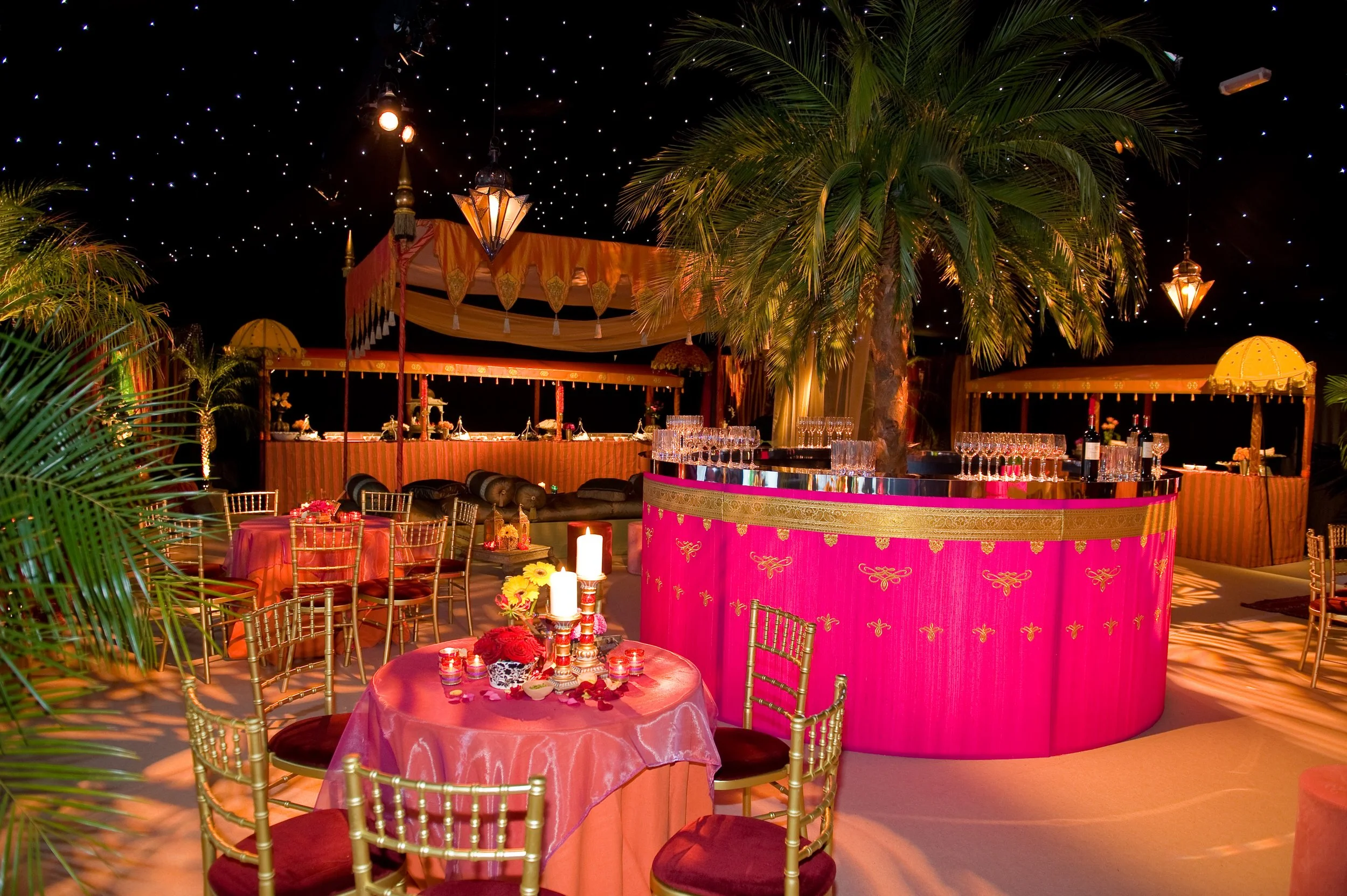 Decorative event space with pink and gold accents, table with candles and flowers, palm trees, and hanging lanterns under a starry ceiling.