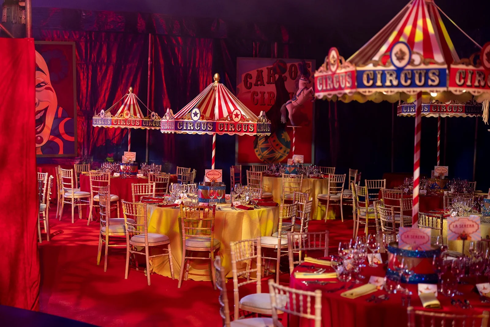 Decorated circus-themed event space with yellow and red tables, chairs, and striped circus tents overhead, featuring colorful banners reading 'CIRCUS' and props like a horse and a clown painting.