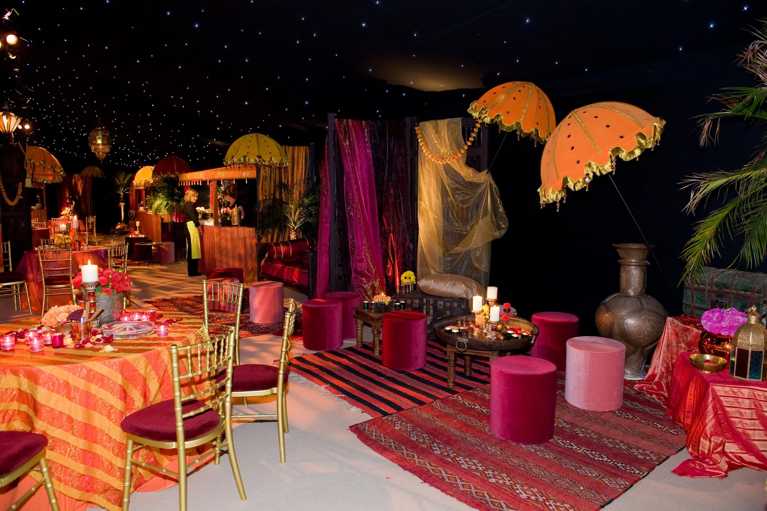 Decorative indoor lounge area with orange umbrellas, candles, flowers, and vibrant drapes, illuminated by star-like ceiling lights.