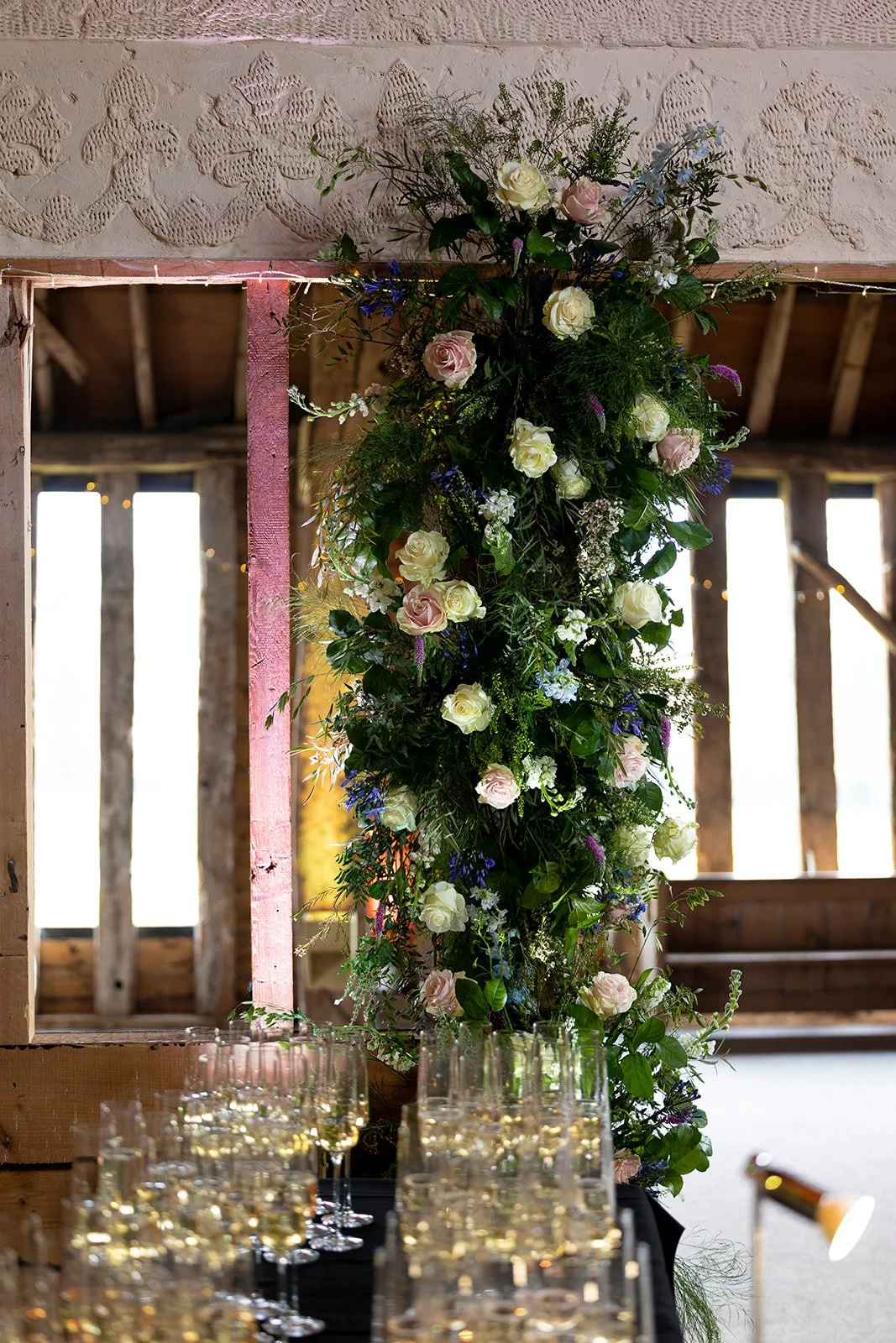 A floral arrangement with white and pink roses, green foliage, and purple and blue accents, set against a rustic wooden barn interior with hanging string lights and a table filled with glasses of champagne.