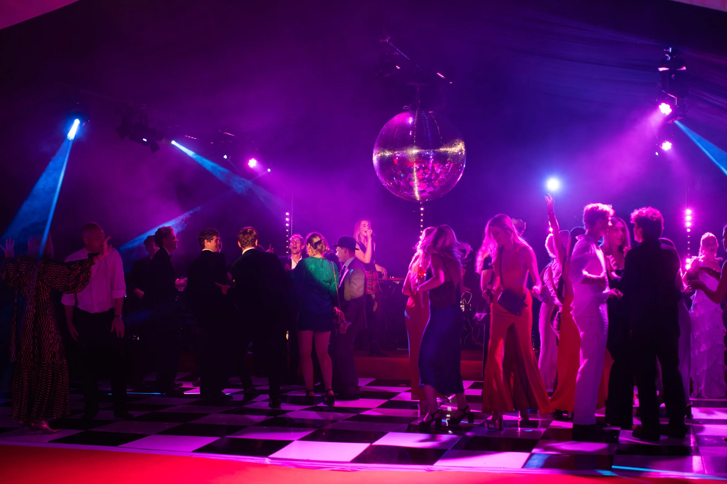 People dancing and socializing on a checkered dance floor under purple and pink lights with a large disco ball overhead.