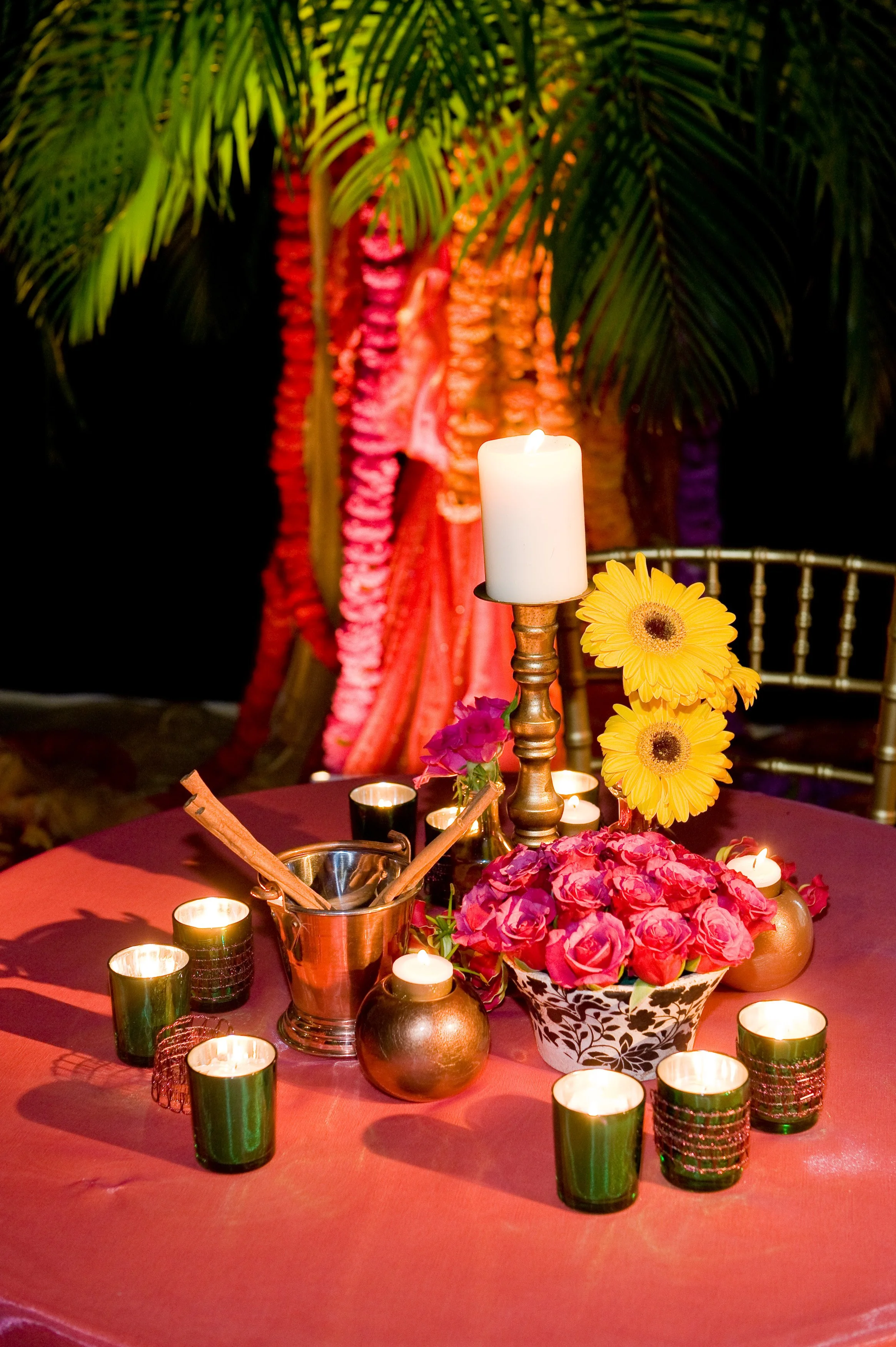 Elegant table centerpiece with white candle, yellow gerbera daisies, pink roses, and small candles, set against a backdrop of green palm leaves and colorful garlands.