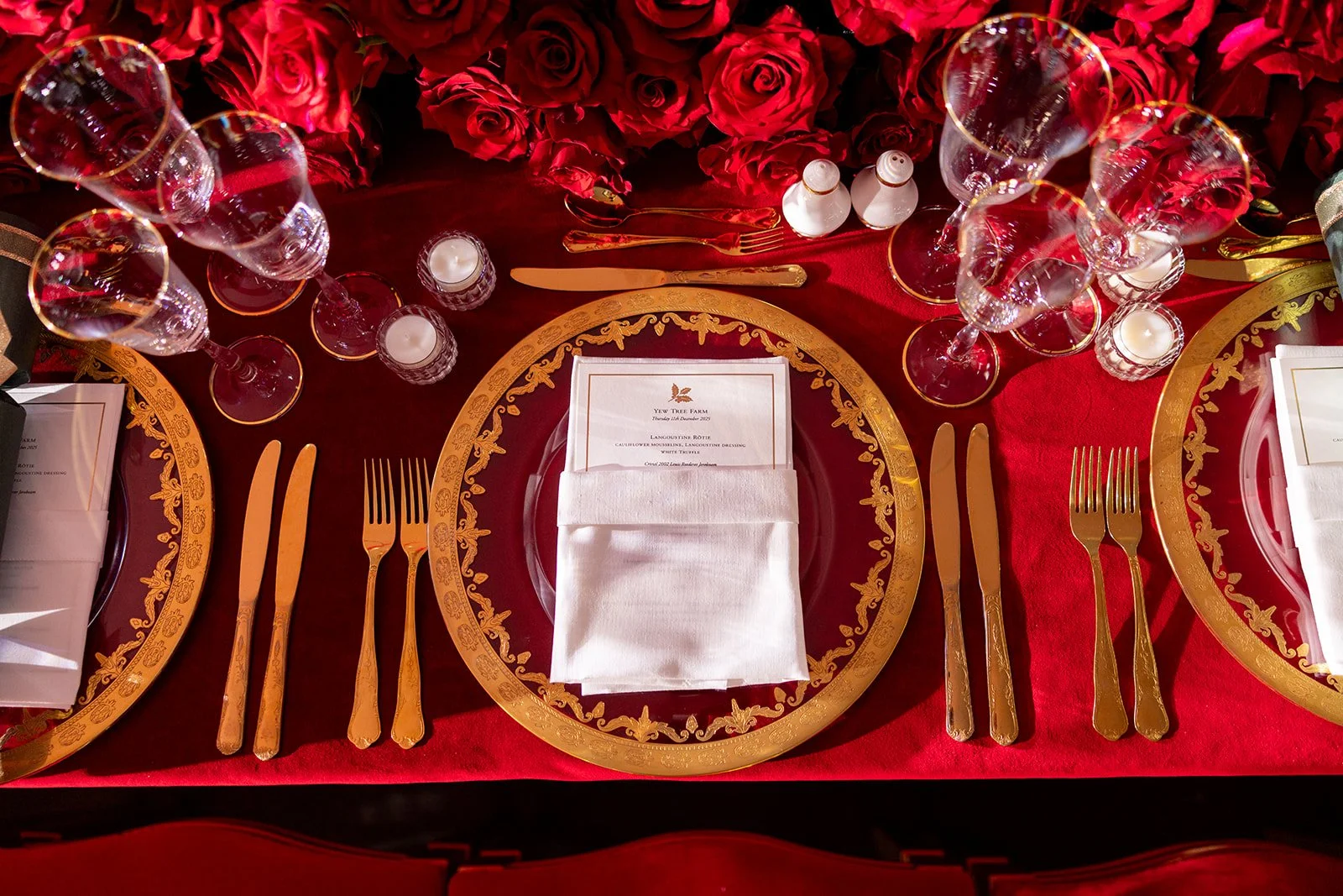 Elegant dining table setting with gold-accented plates, gold cutlery, multiple wine glasses, white napkins, candles, and a large bouquet of red roses in the background.