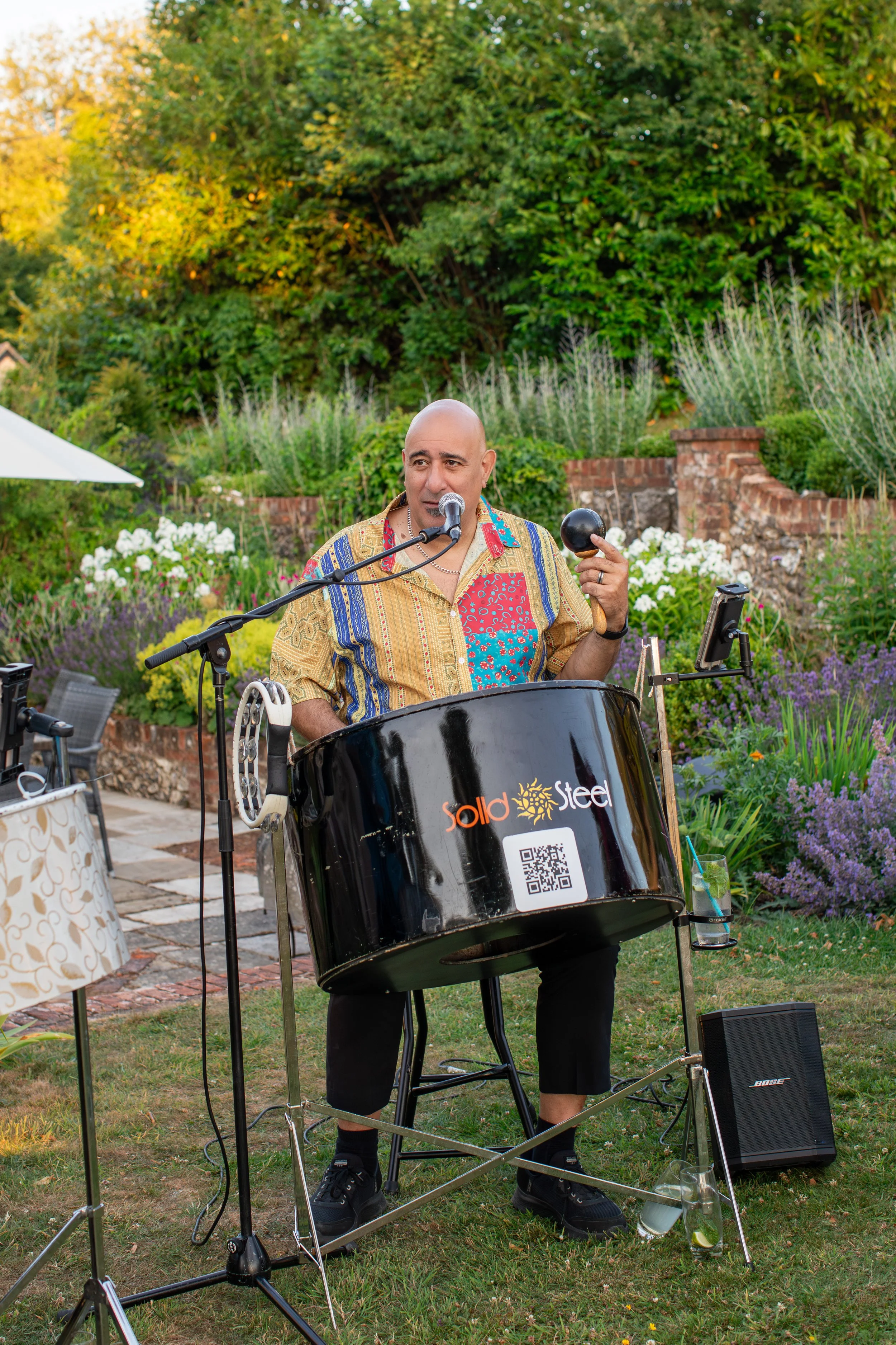 A man with a shaved head playing percussion instruments outdoors in a garden setting, with greenery and flowers in the background.