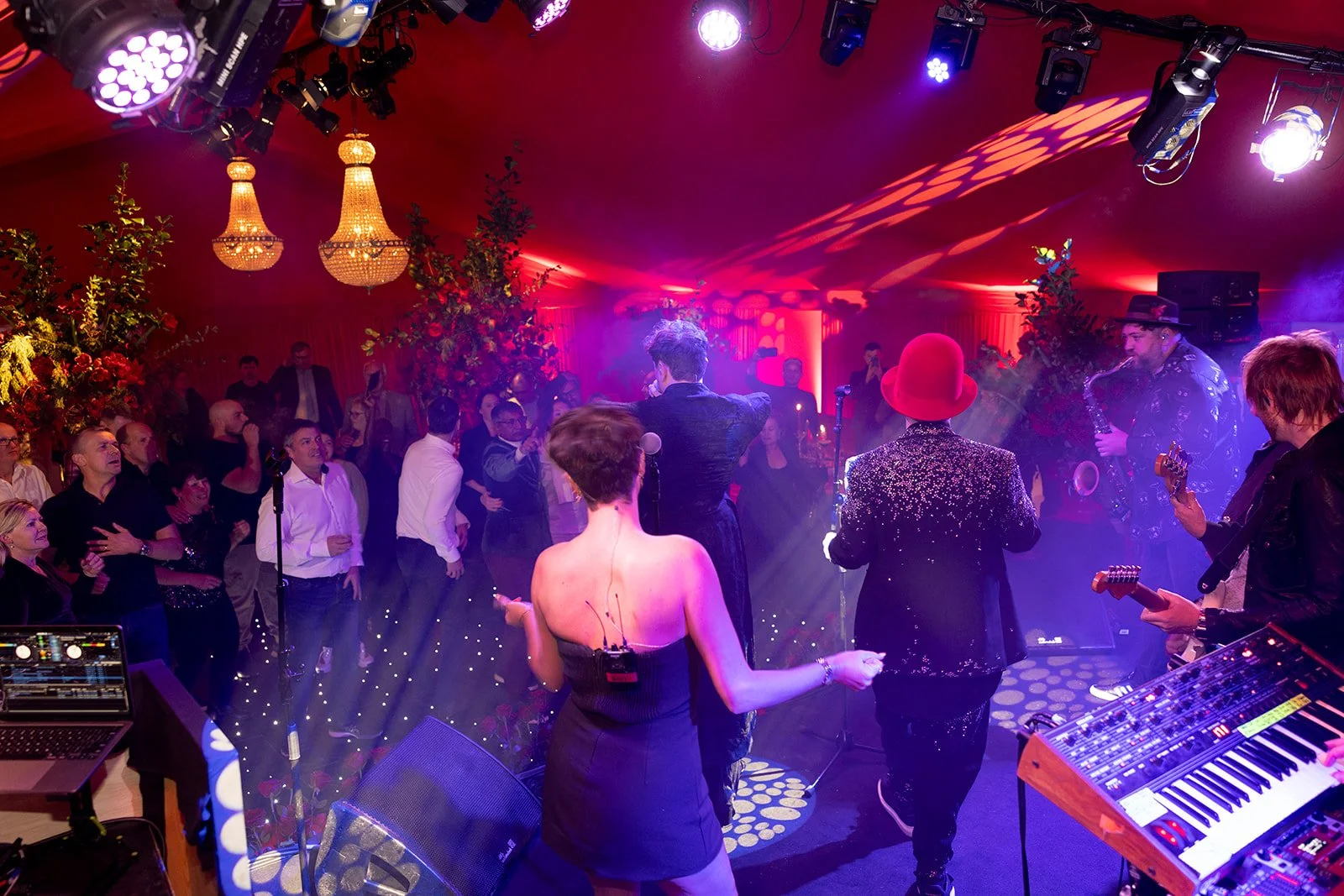 A lively party scene with a band performing on stage, surrounded by guests dancing and enjoying the music in an elegantly decorated venue with chandeliers and floral arrangements.
