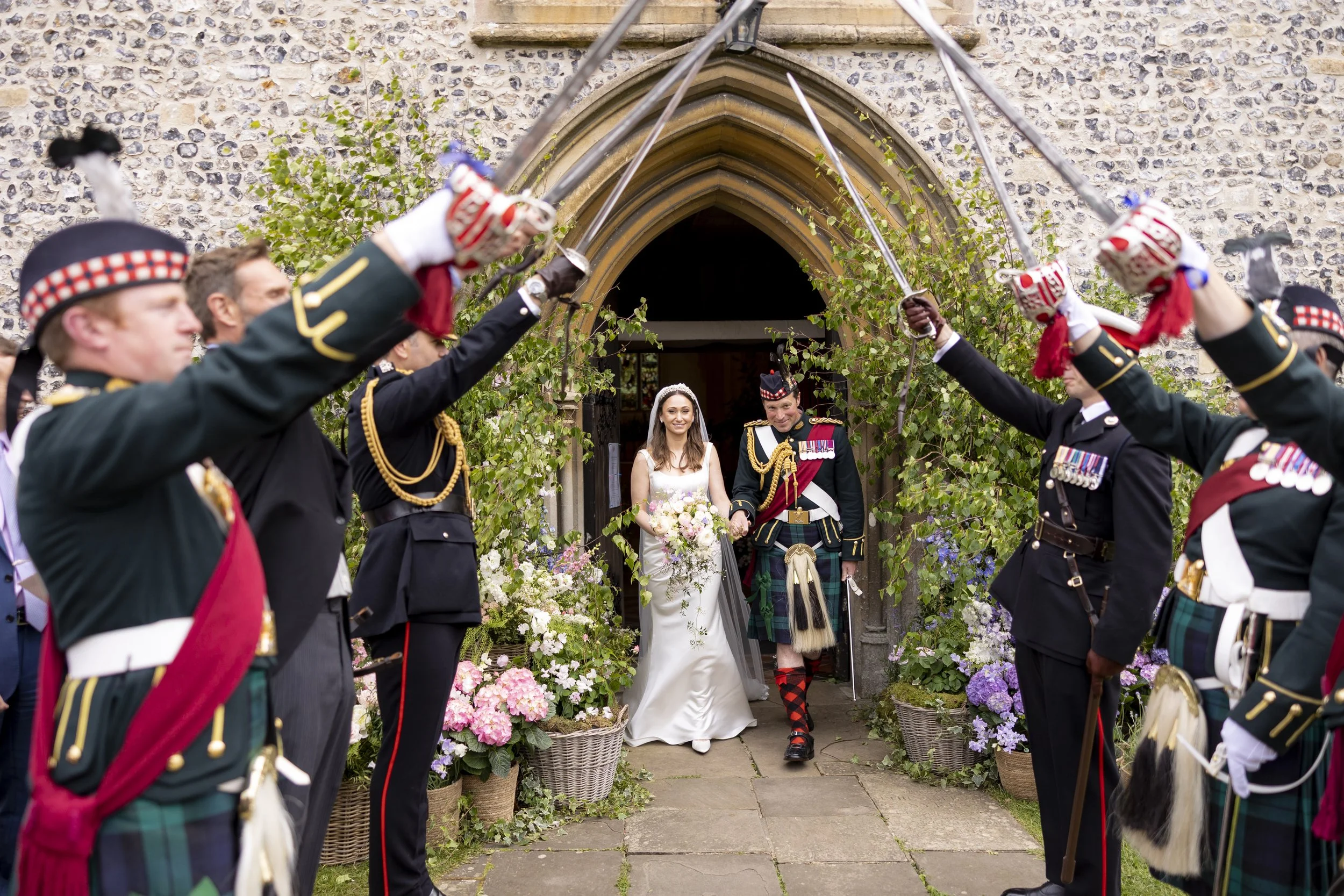 A bride and groom walking through a flower arch, surrounded by uniformed soldiers holding swords creating an arch of swords. The scene is outside a stone building with an arched wooden door, decorated with flowers and greenery.