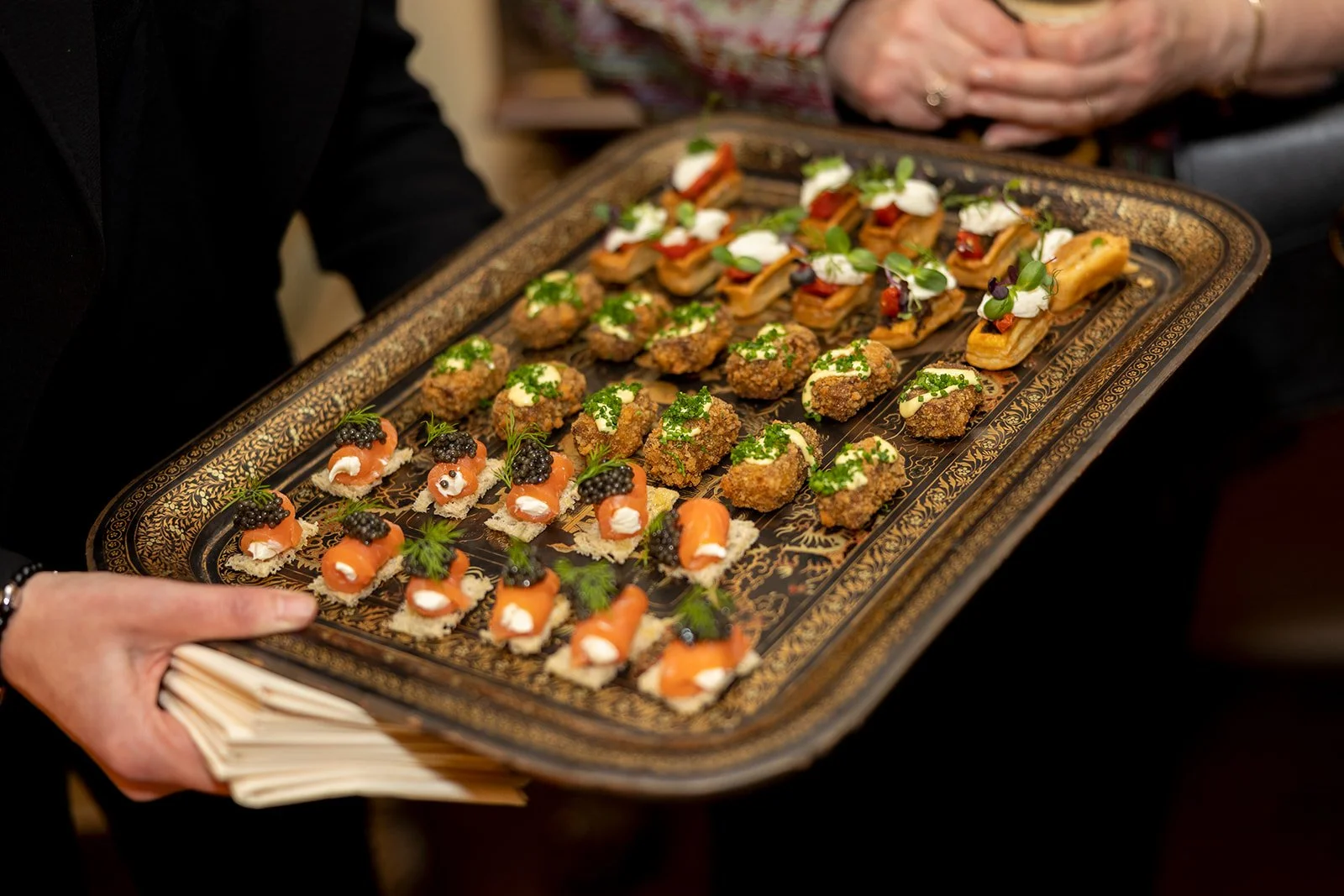 A person holding a large ornate tray with an assortment of small fancy hors d'oeuvres, including smoked salmon with cream cheese and black caviar, fried bites with chopped herbs, and small canapé sandwiches garnished with microgreens and dollops of c