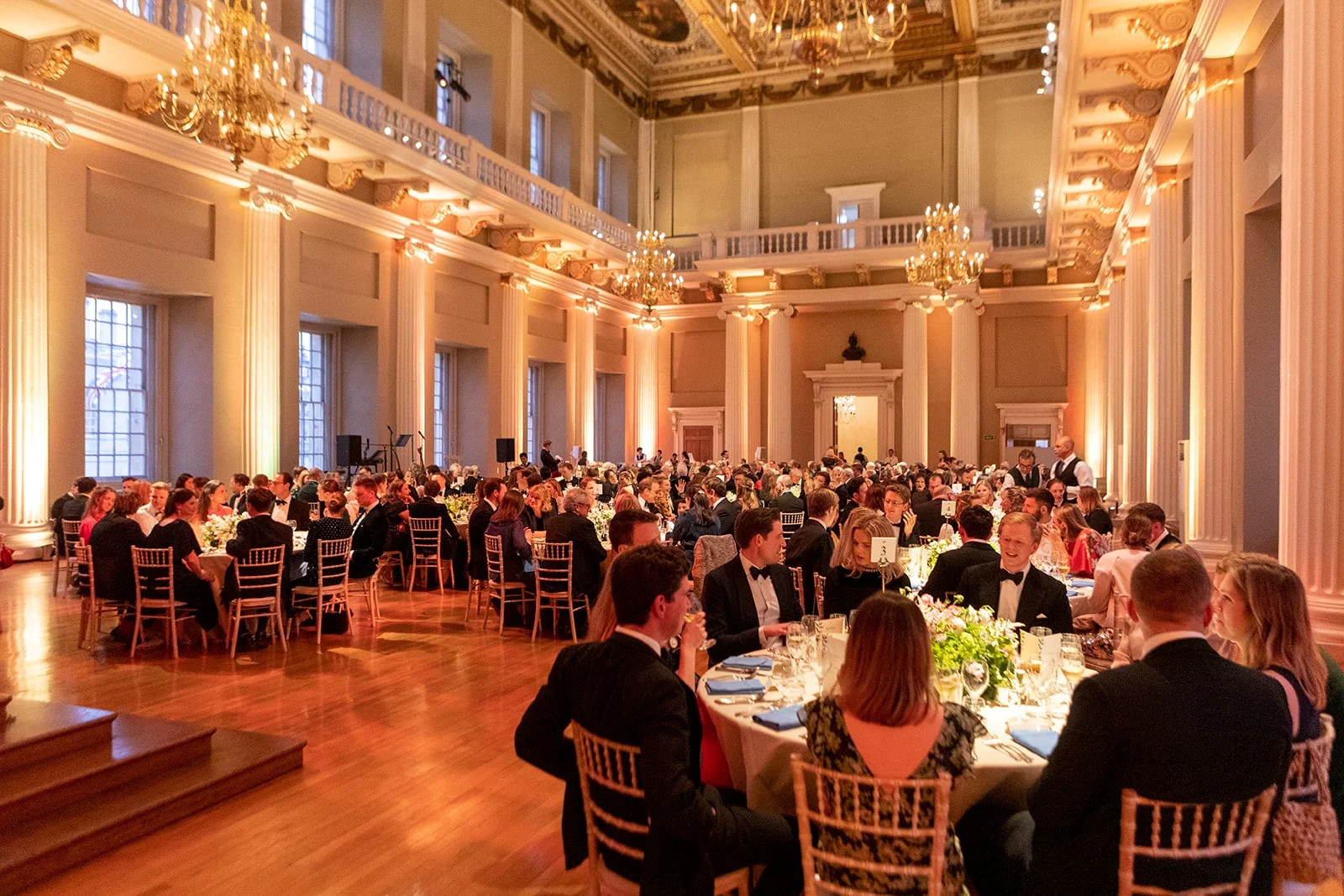 A large formal banquet hall filled with elegantly dressed guests seated at round tables decorated with floral centerpieces. The hall features high ceilings, tall windows, ornate chandeliers, and classical architecture with columns.