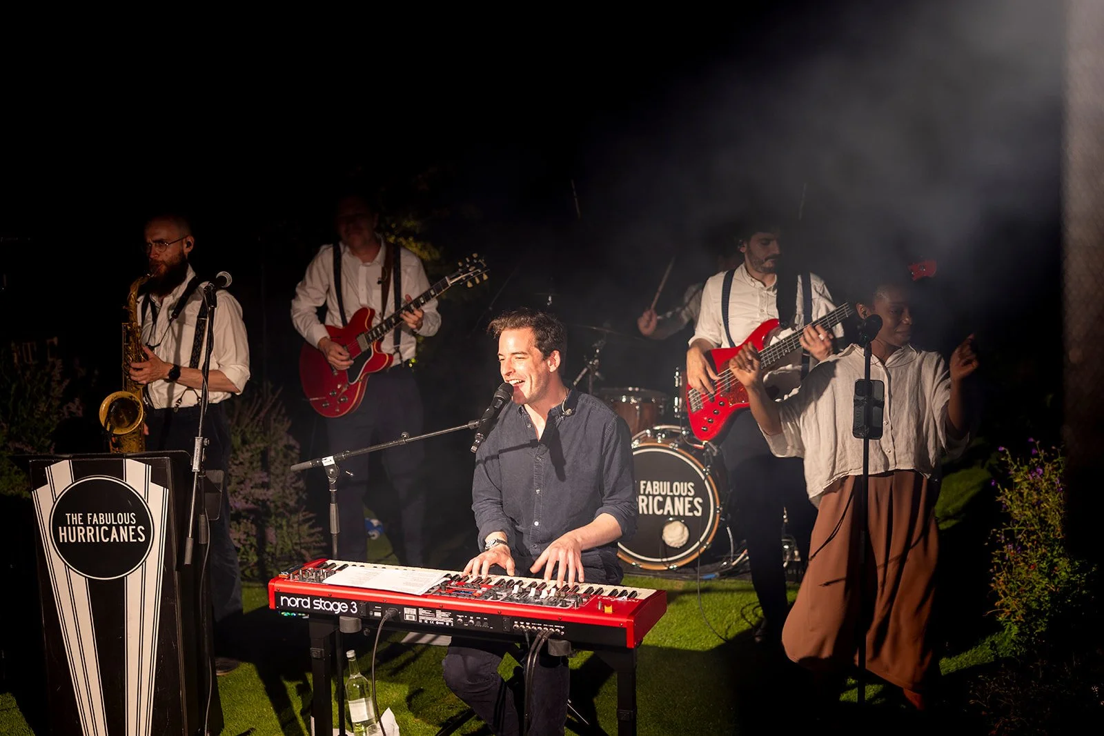 A live musical performance featuring a band called The Fabulous Hurricanes. The band includes a keyboard player, two guitarists, a drummer, a saxophonist, and a singer, all performing outdoors at night.