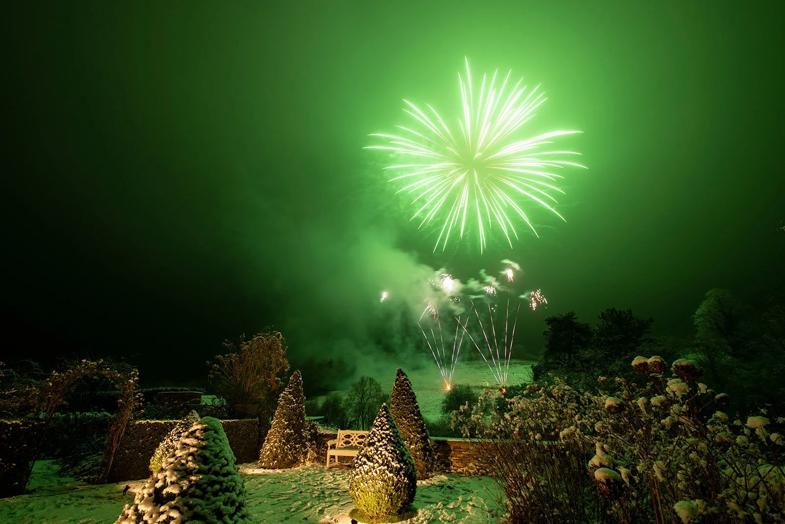 Fireworks display illuminating a snow-covered garden at night.