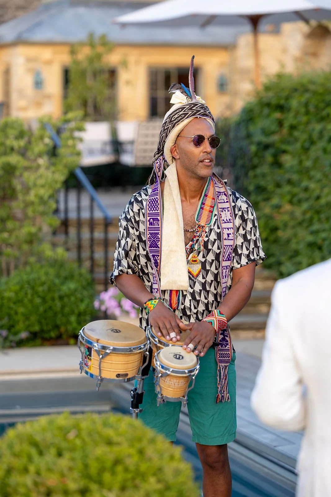 A man playing drums outdoors, wearing sunglasses, a patterned shirt, colorful beaded jewelry, and a feathered headpiece.