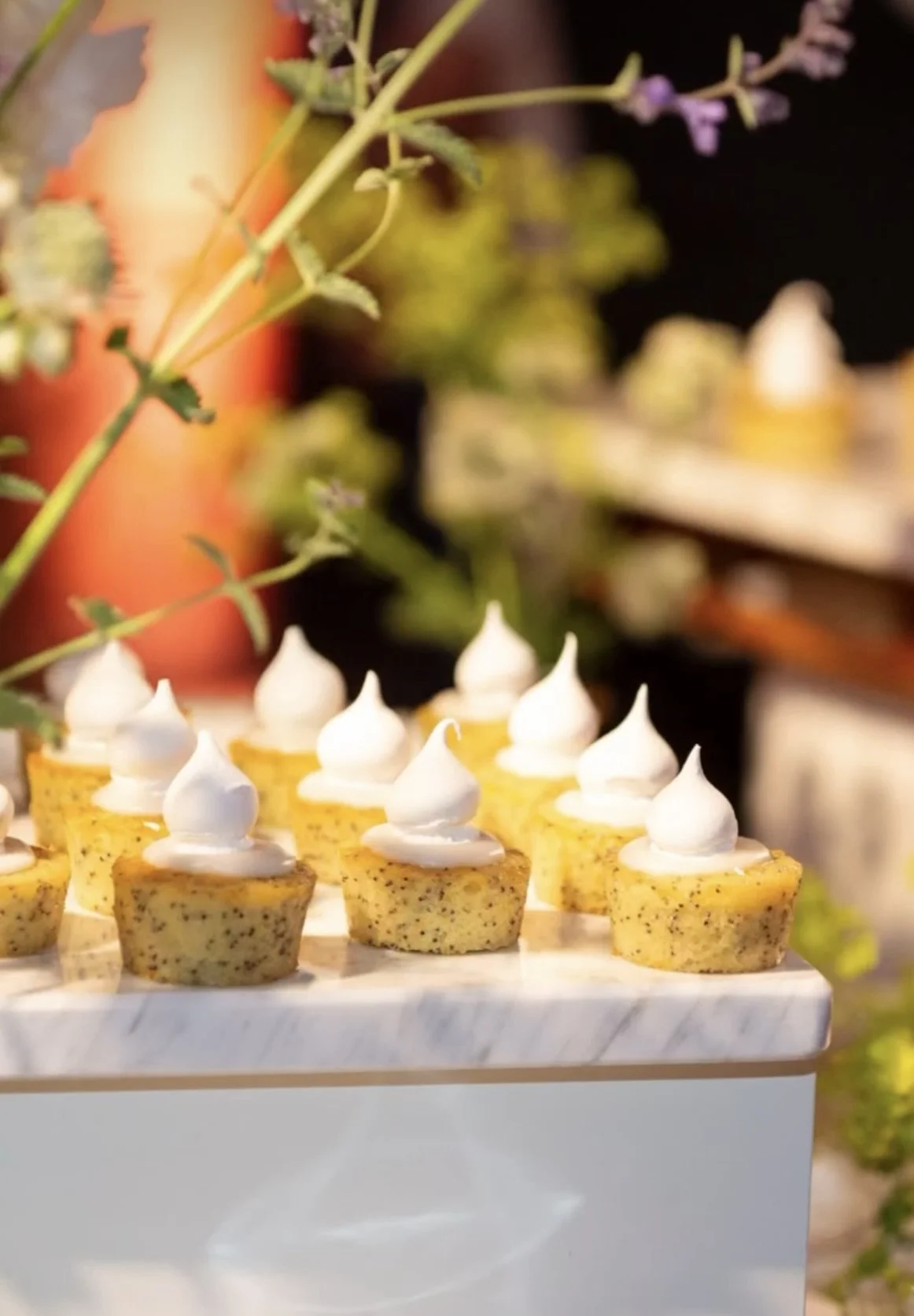 Miniature yellow cupcakes topped with white frosting, displayed on a marble surface with flowers in the blurred background.