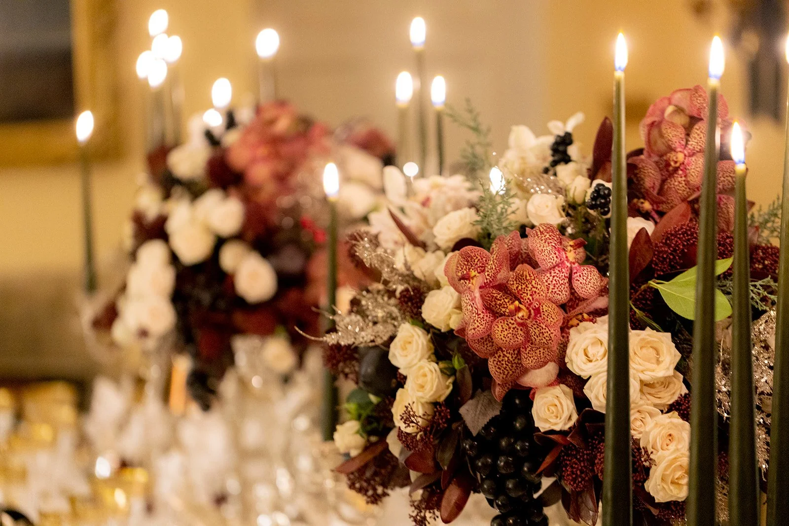 A floral arrangement featuring white roses, orange orchids, black berries, and greenery, with lit candles in the background.
