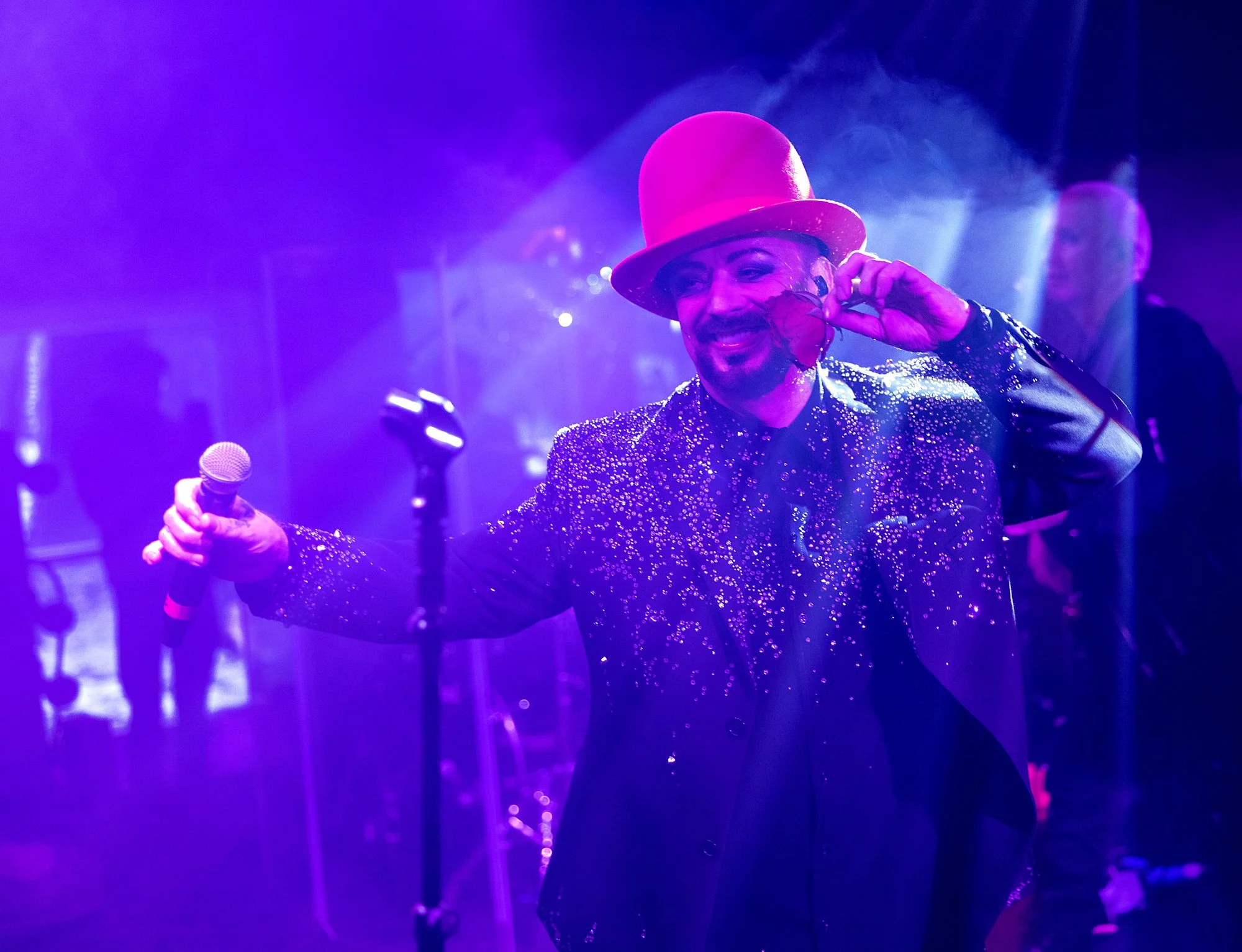A man in a sparkling black jacket and pink hat smiling and holding a red rose in a nightclub with purple lighting.