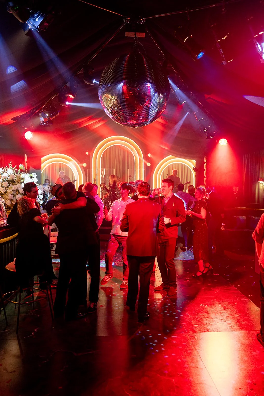 People dancing and socializing in a nightclub with colorful lighting, a disco ball hanging from the ceiling, and neon arches in the background.