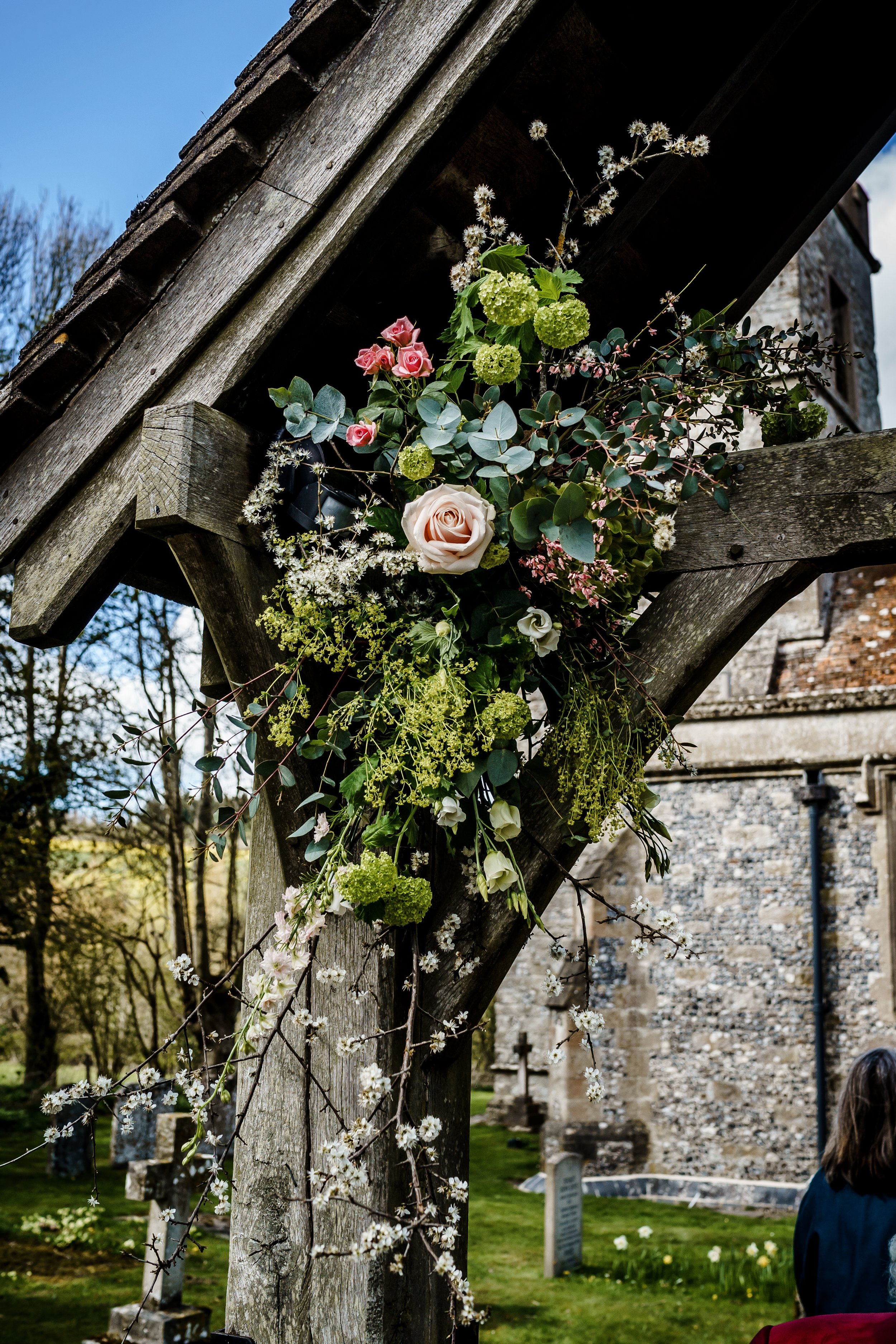 A wooden archway decorated with various flowers and green foliage, situated outdoors near a stone building with a graveyard in the background.