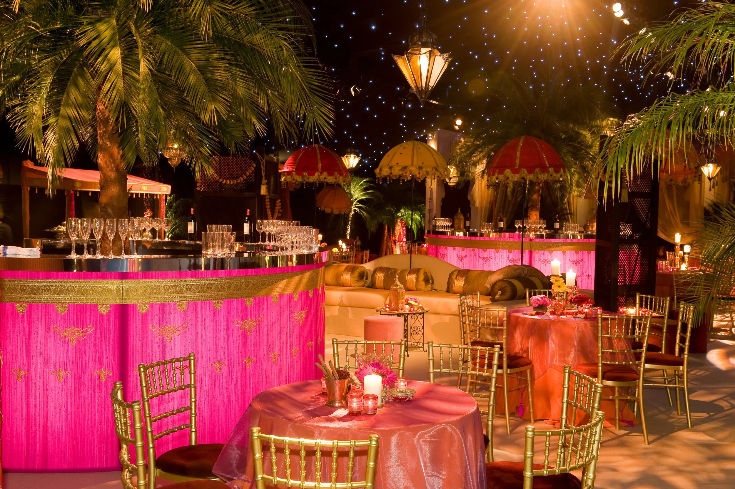 Decorative outdoor dining or event setup at night with pink fabric-wrapped bar, gold chairs, tables with pink tablecloths, lit candles, umbrellas, palm trees, and starry string lights overhead.