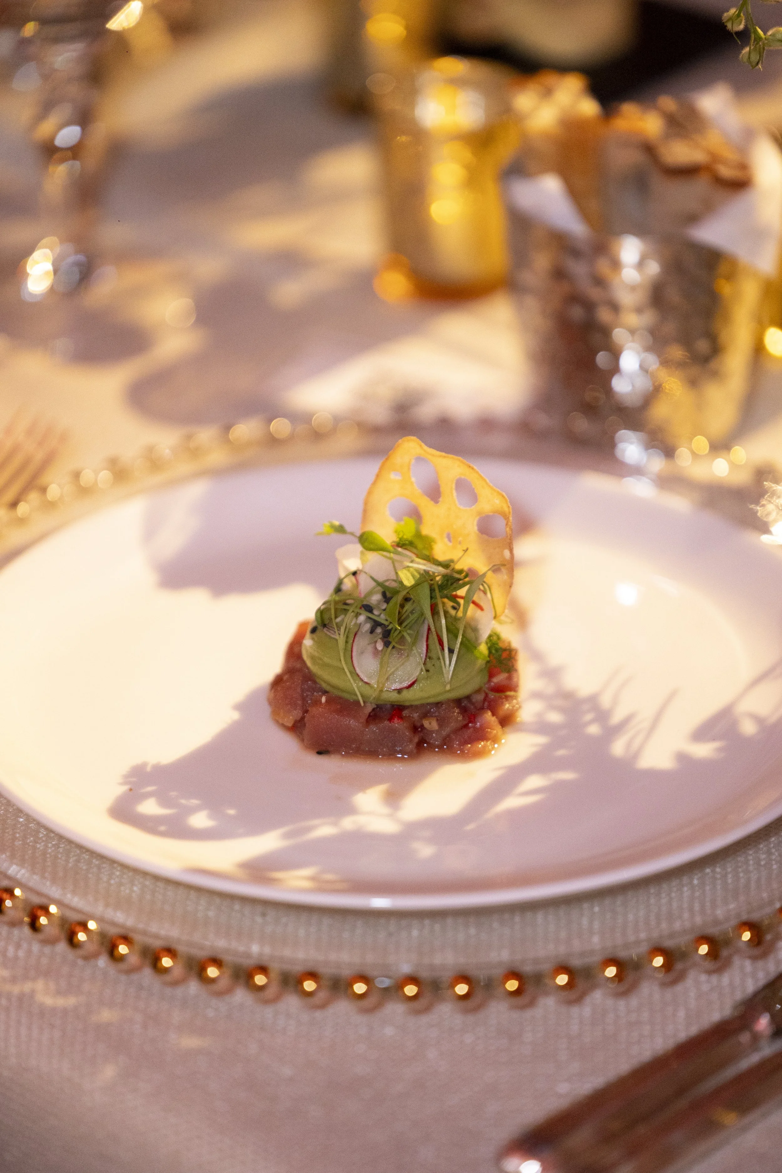A gourmet appetizer consisting of a small piece of beef topped with avocado, microgreens, and a thin crispy lotus root chip, served on a white plate with a decorated gold and pearl bordered charger under it.