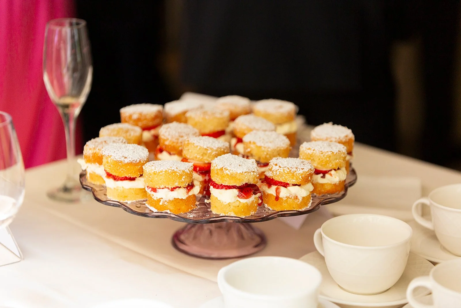 A glass cake stand with mini strawberry shortcakes topped with powdered sugar on a table set for a gathering.