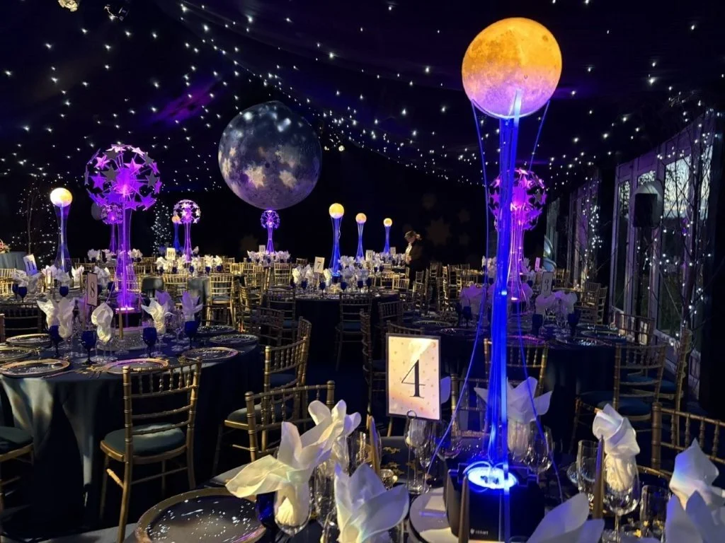 Indoor event decorated with moon and star themed decor, illuminated sphere centerpieces, and ceiling lights resembling stars, suggesting a space or celestial themed celebration or party.