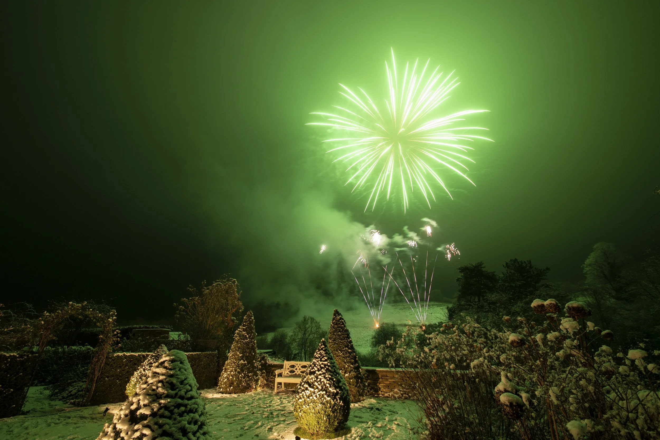 Fireworks display lighting up the night sky with green hues, seen from a snowy landscape with decorated trees and a park bench.