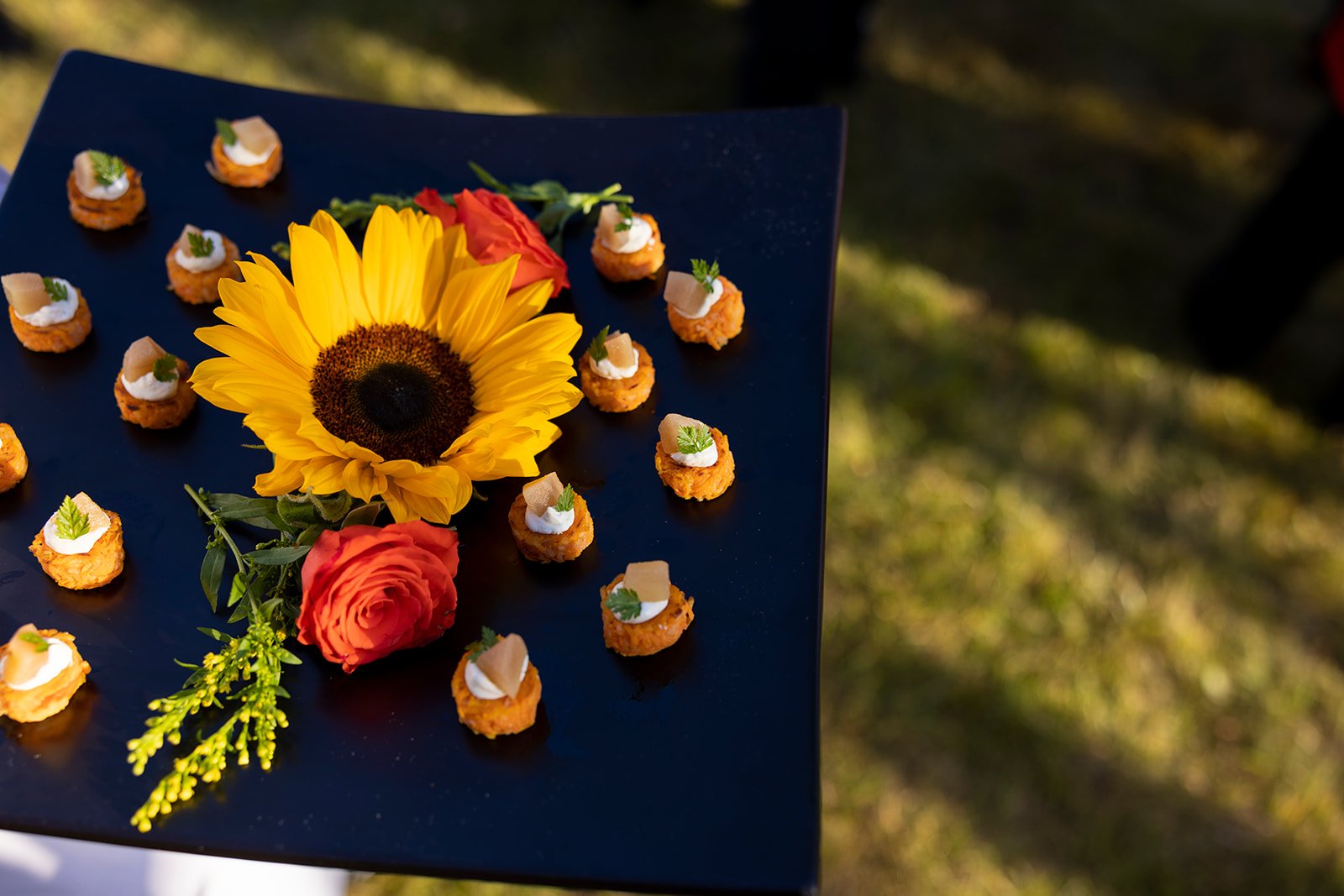 A black tray with small appetizers topped with white sauce and herbs, accompanied by a sunflower, a pink rose, and greenery, set outdoors on a sunny day.
