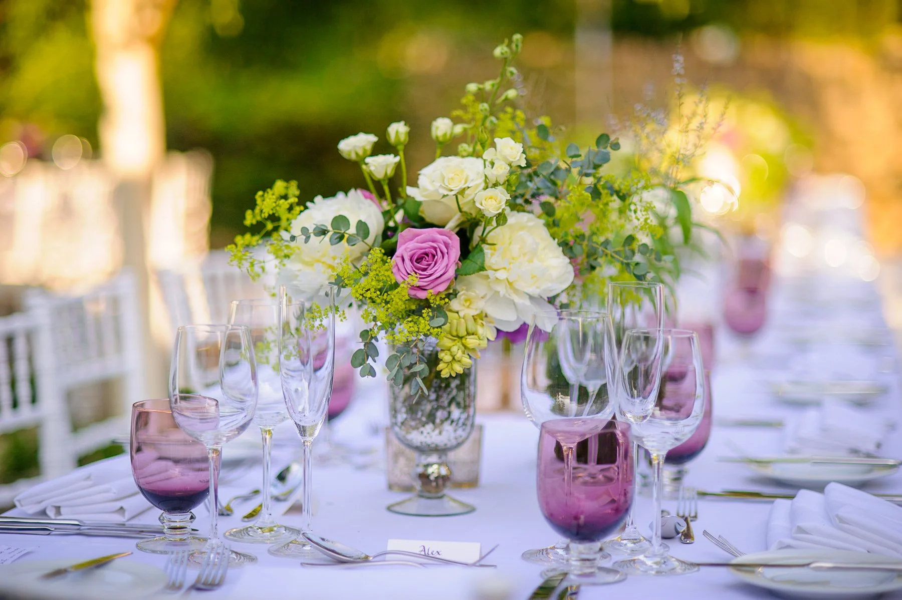 A beautifully arranged outdoor wedding or event table with a floral centerpiece featuring white and purple roses and greenery, surrounded by wine glasses and tableware on a white tablecloth.