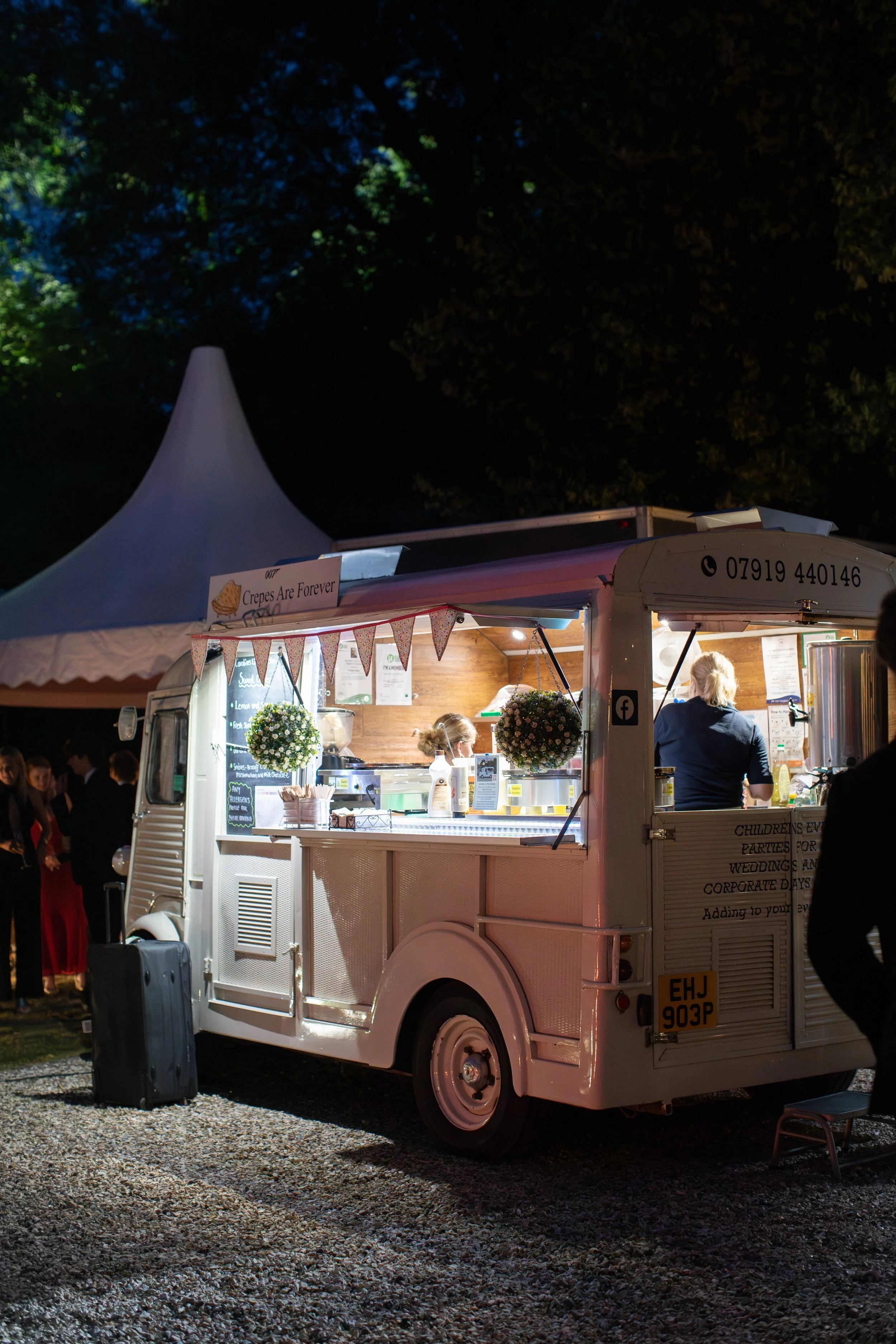 Night scene of a white food truck selling crepes, with a sign that reads "Crepes Are Forever." The truck is decorated with bunting and hanging flower baskets, sitting on gravel with people nearby and a tall white tent in the background.