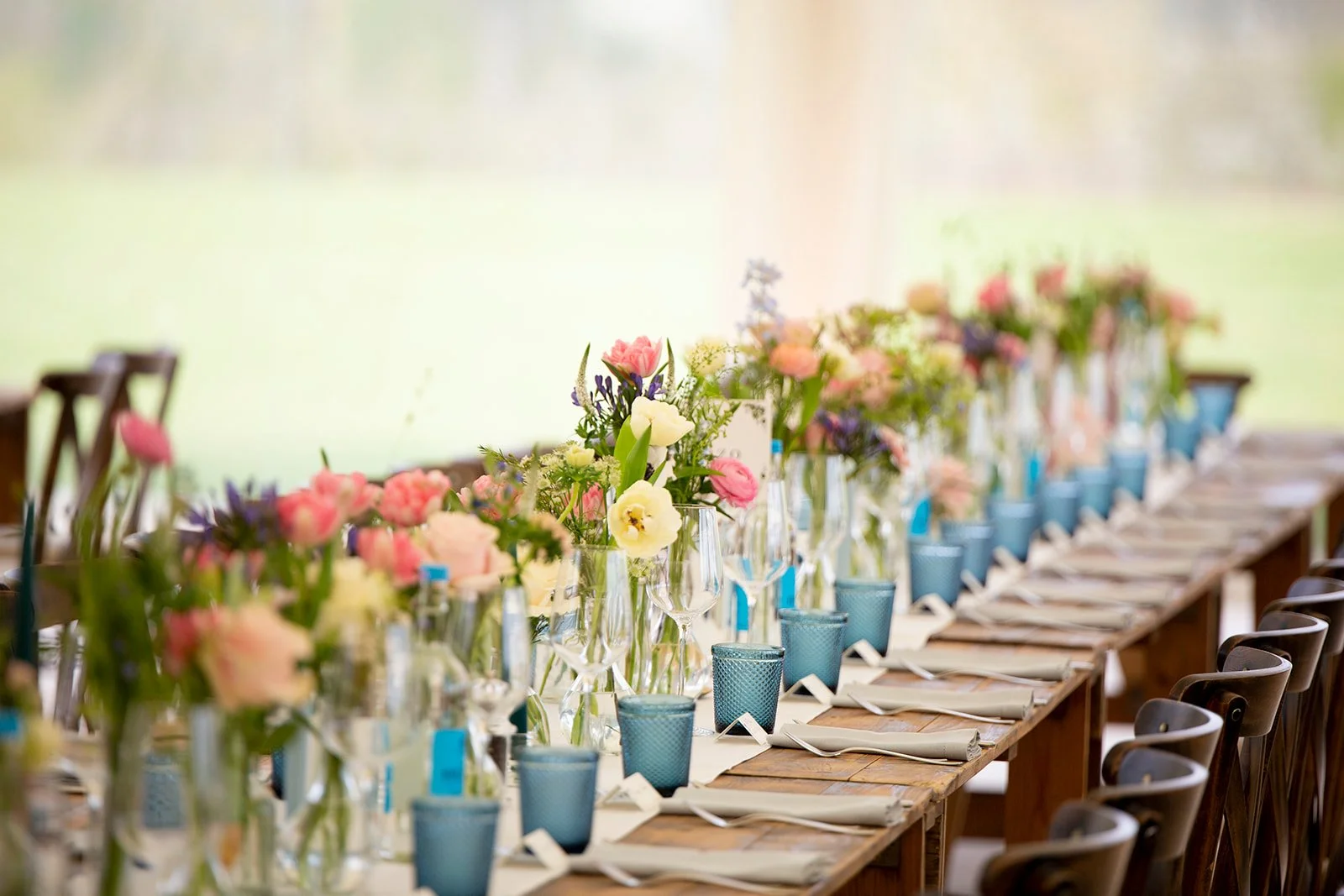 A long table decorated with pink, purple, and white floral centerpieces, set for a formal event with menus, blue cups, and napkins.