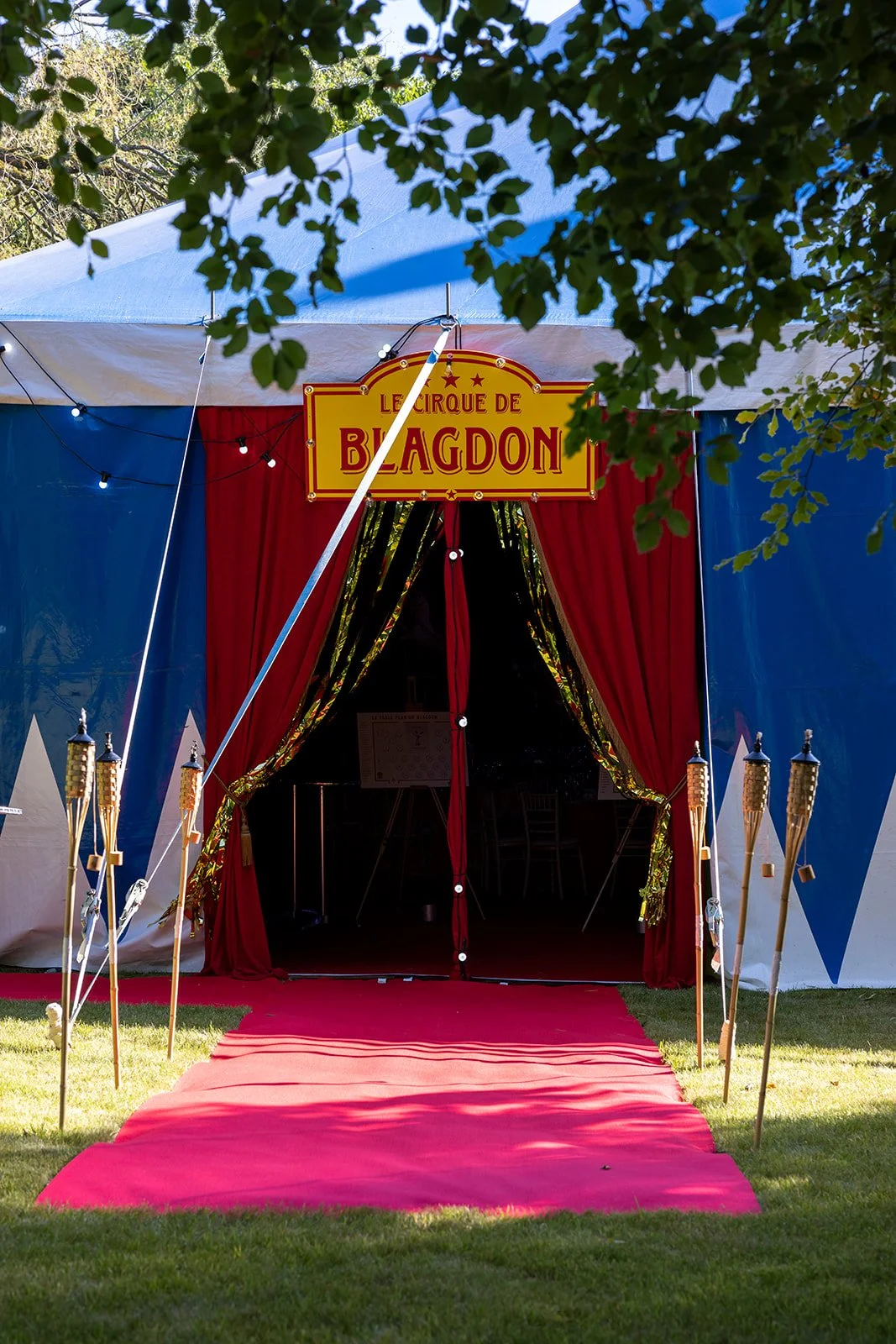 Entrance to a circus tent with red curtains and a yellow sign that reads "Le Cirque de Blagdón." There are tiki torches on both sides of the entrance and a red carpet leading inside. Green foliage is visible at the top of the image.