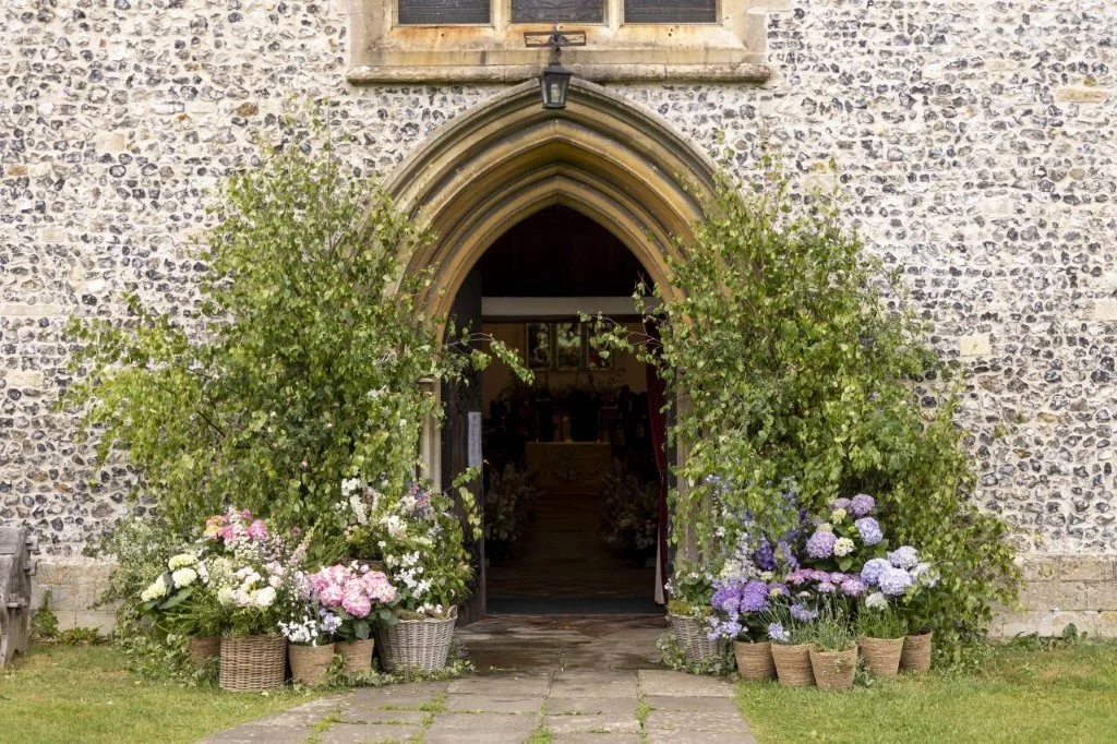 Stone church entrance with an arched doorway framed by green bushes and flower pots of white, pink, purple, and blue flowers.