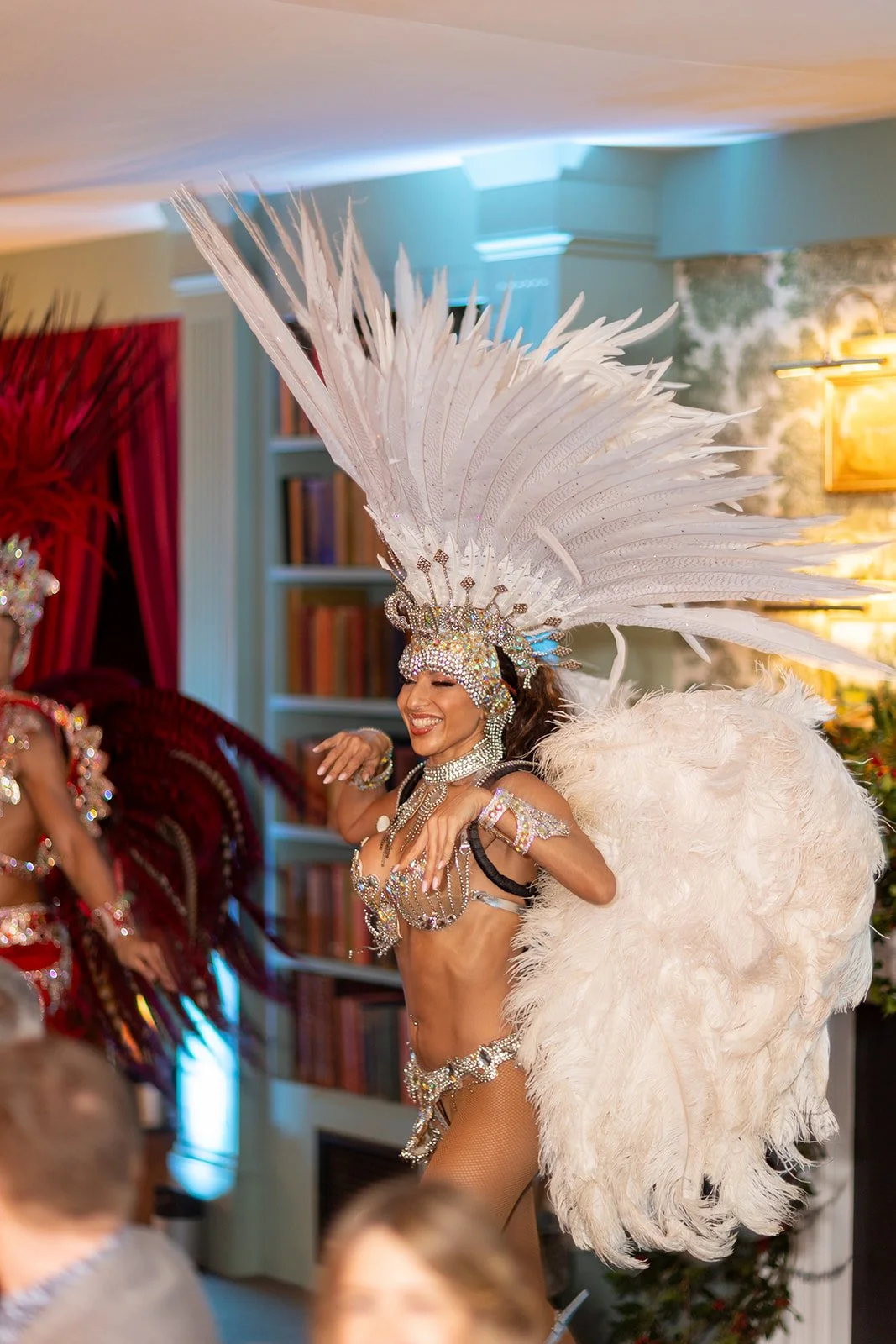 A woman in a colorful costume with feathers, jewelry, and a headdress dancing at an indoor event.