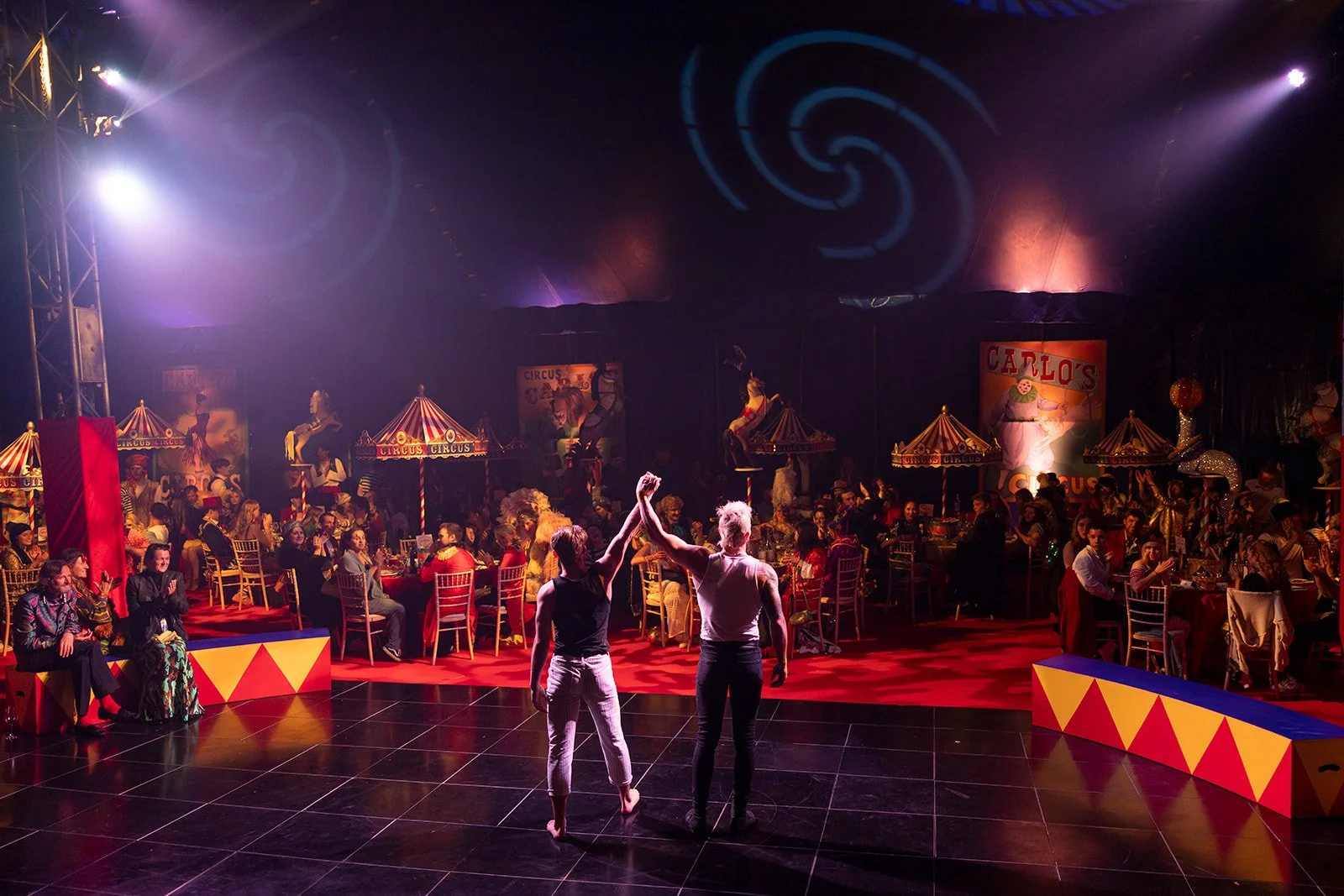 A circus-themed performance with two performers holding hands in the center, surrounded by an audience seated at decorated tables, with circus props and figures in the background under colorful lighting.