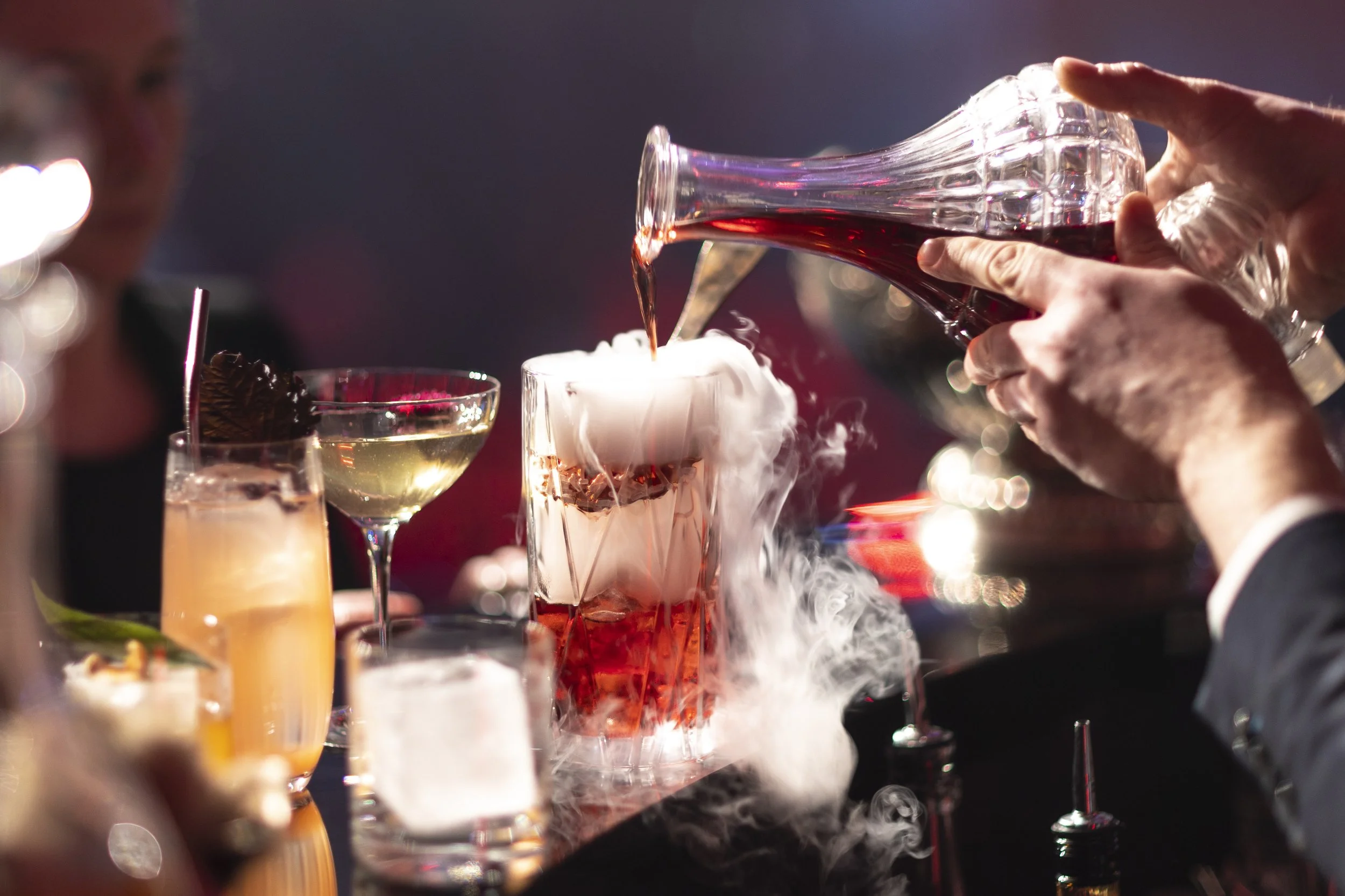 A person is pouring red wine into a glass with dry ice, creating smoke and fog effects on a bar or party setting.