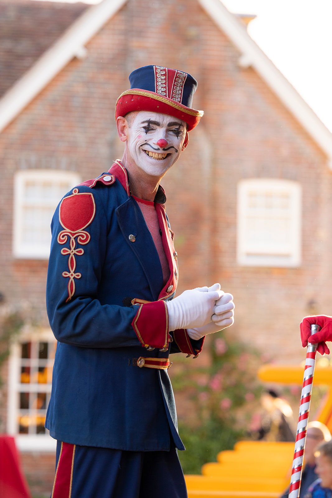 A smiling man dressed as a circus clown in a colorful blue and red costume with a hat, white face paint, and a red nose, standing outdoors in front of a brick building.