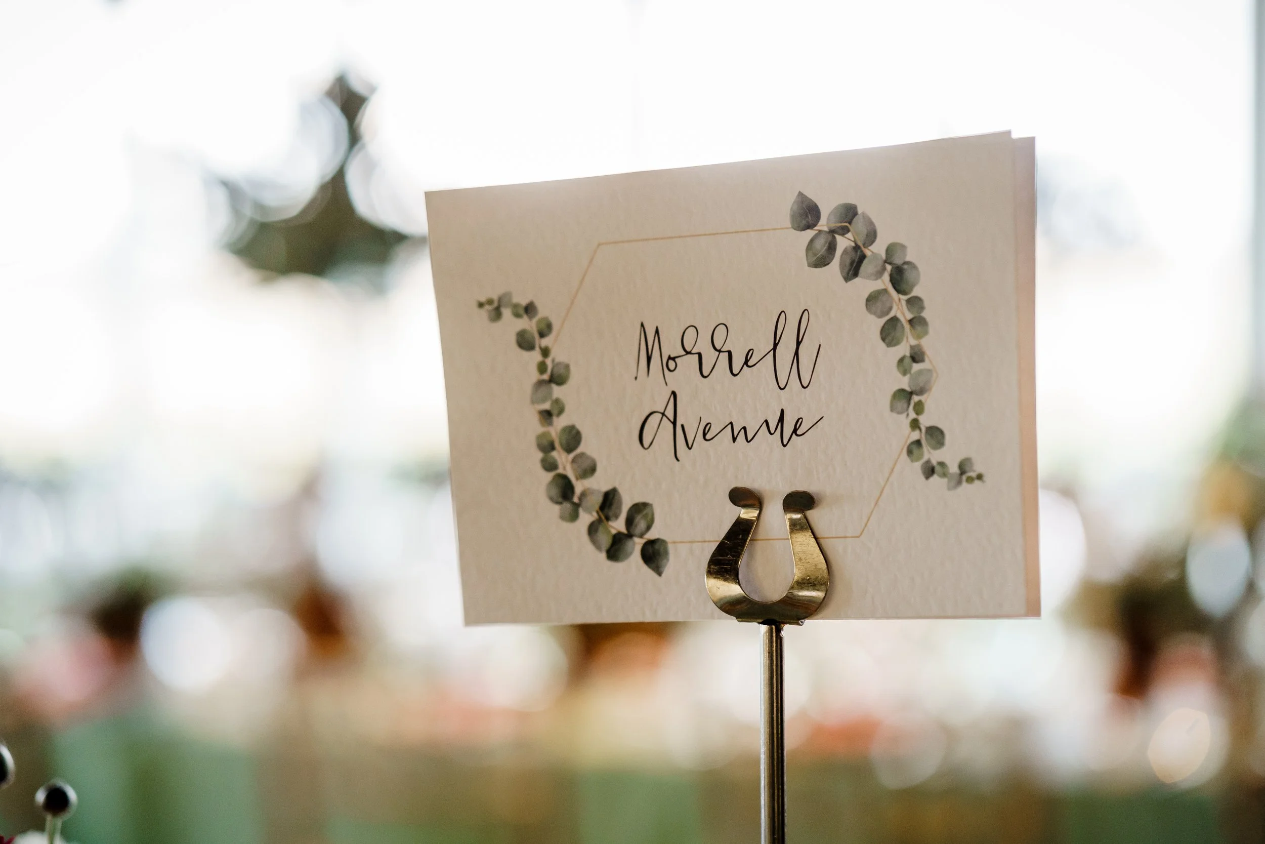 A wedding or event table number card with the name 'Morrett Avenue' and a botanical wreath border, attached to a metal holder with a gold number 2.