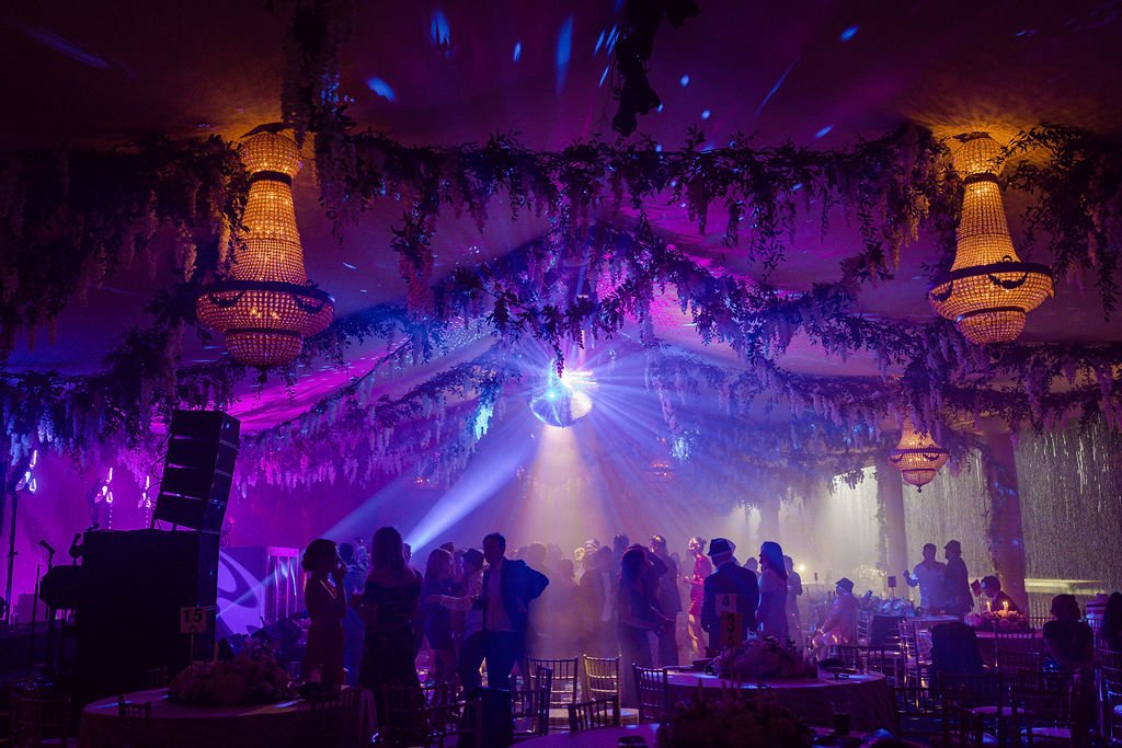 People dancing and socializing in a decorated event hall with purple lighting, chandeliers, and a disco ball.