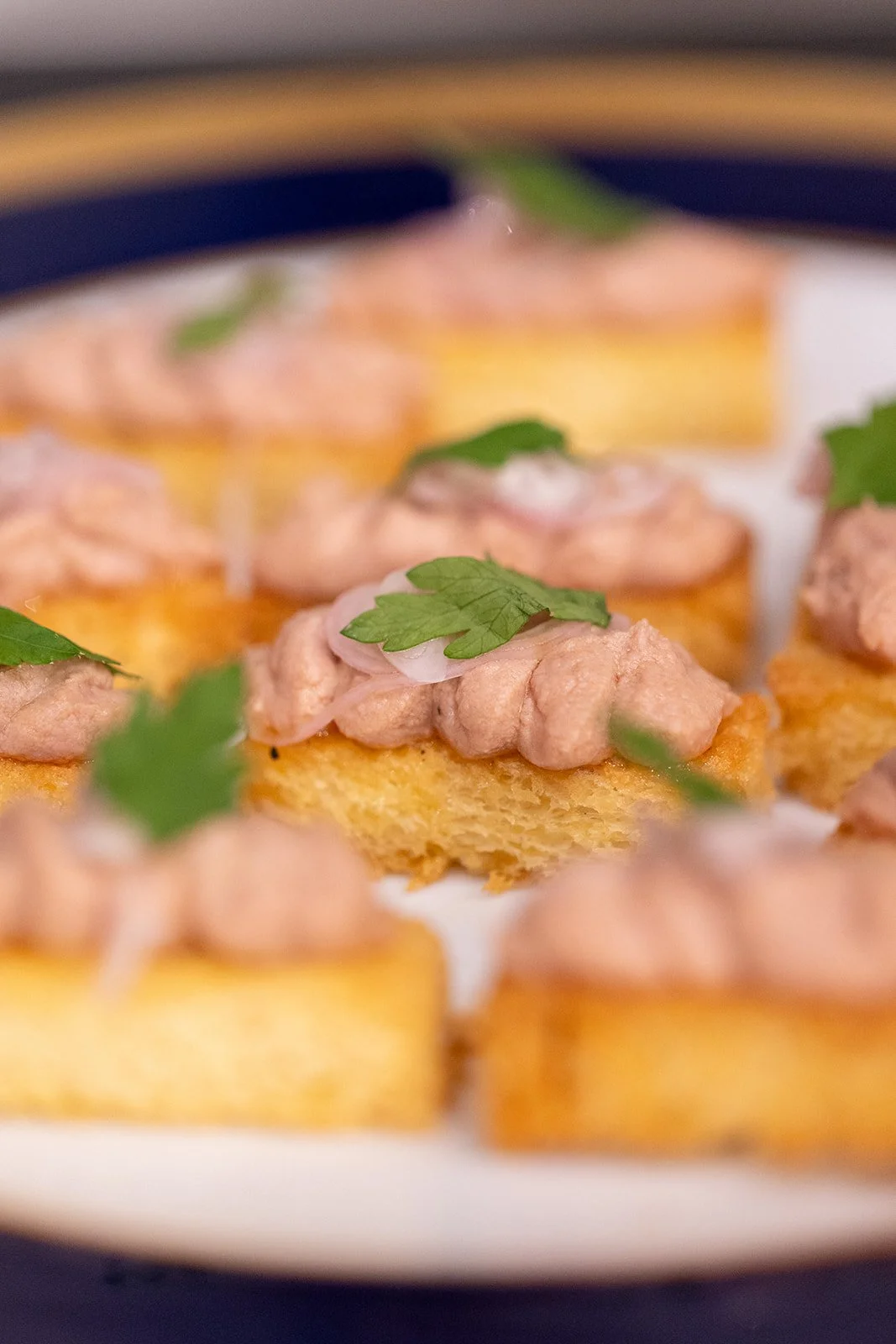 Close-up of canapé bites with cream-colored toast, topped with pinkish meat spread and fresh green parsley leaves.