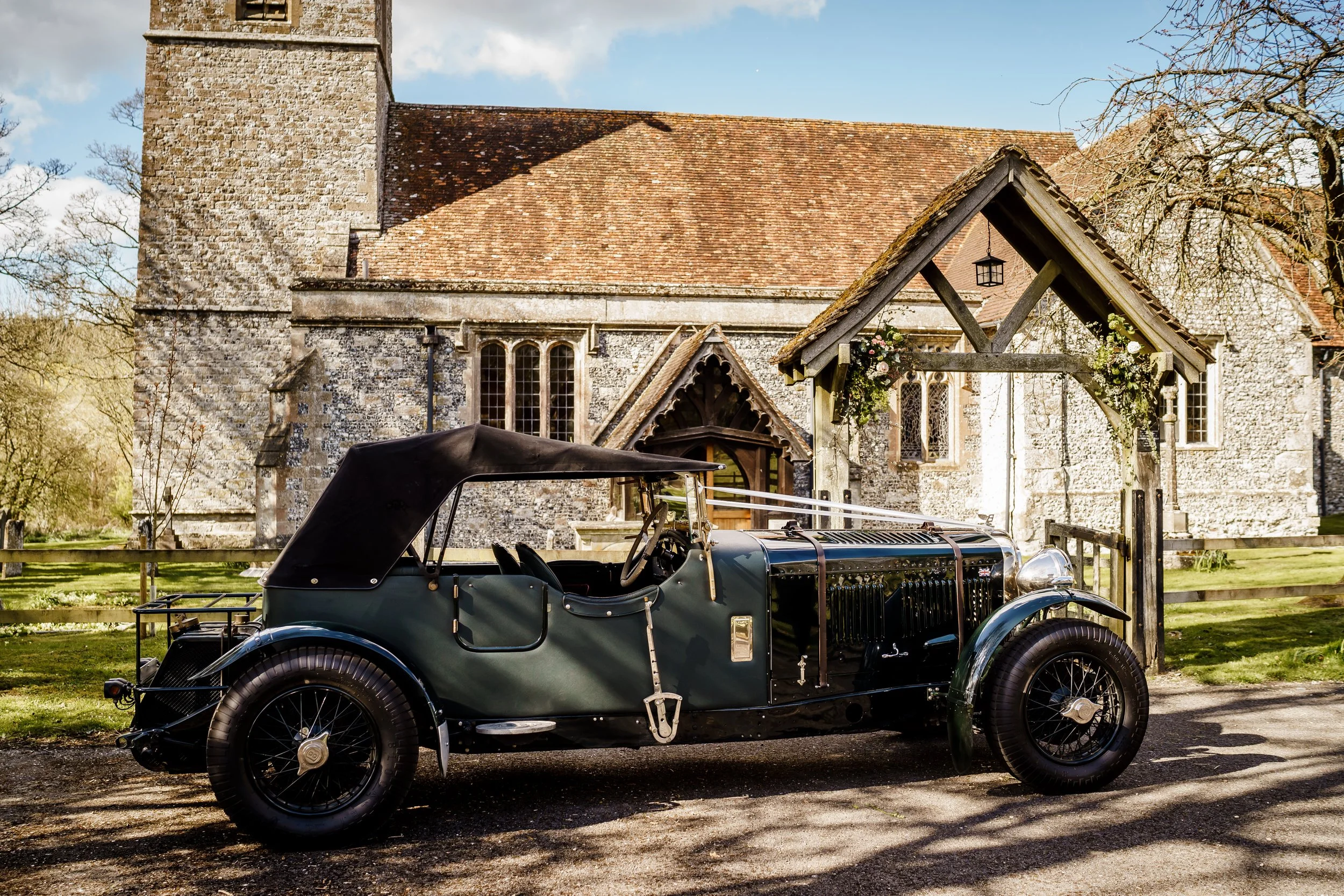 An antique black vintage car parked in front of an old stone church with a sloped roof and a clock tower, surrounded by trees and a grassy area.