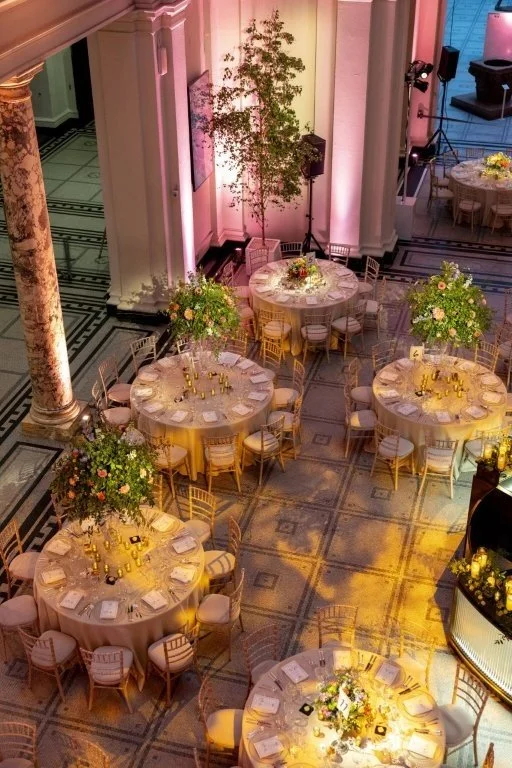 Elegant event setup with round tables, floral arrangements, and gold chairs in a grand hall.