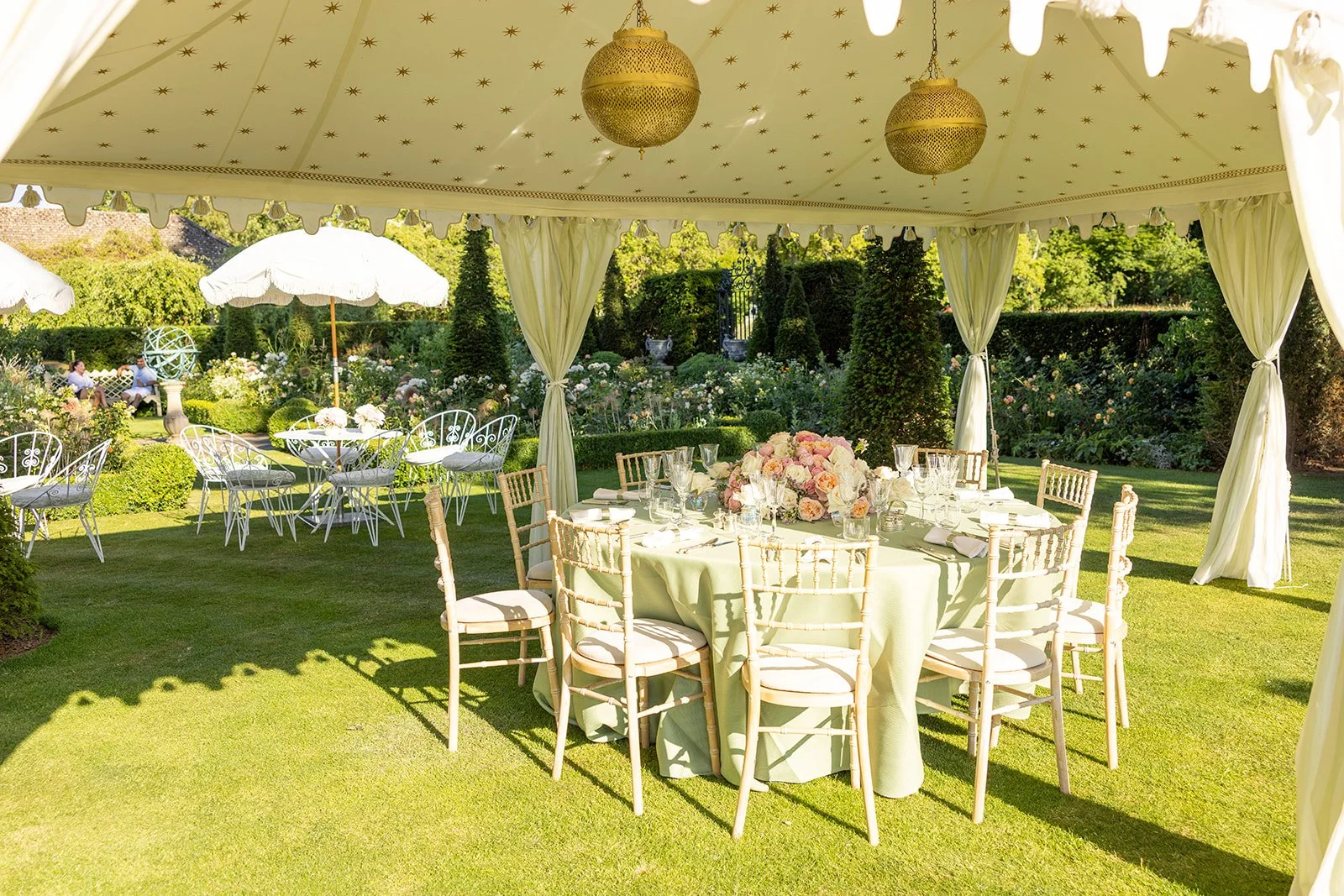 An outdoor garden setting with a tent structure, decorated for a celebration, featuring a round table with a floral centerpiece, surrounded by wooden chairs, and additional seating with chairs and parasols in the background.