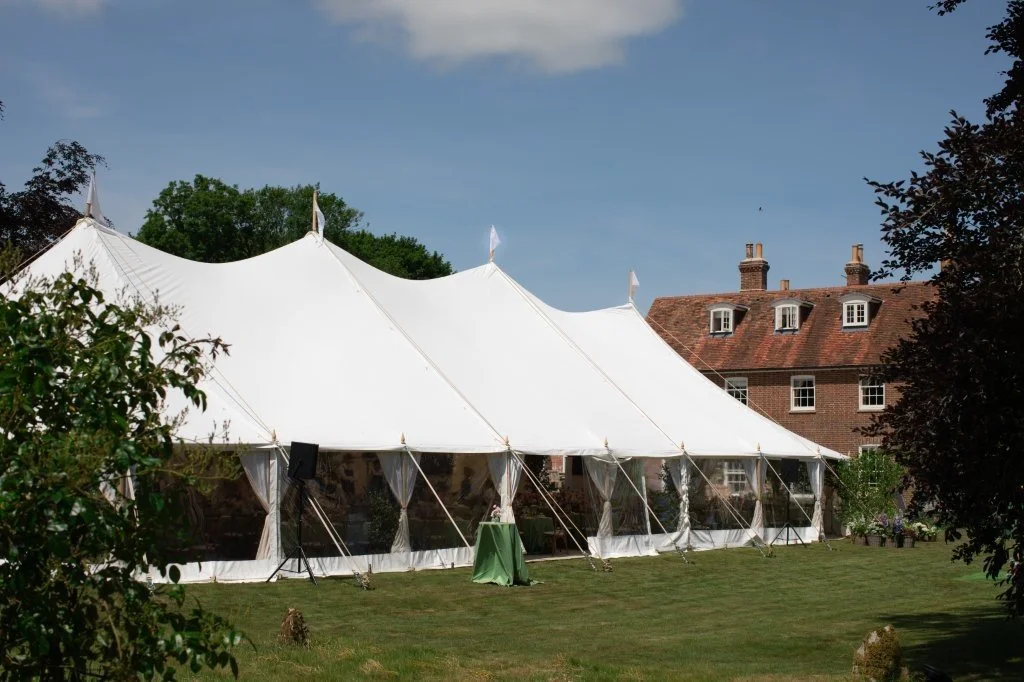 A large white event tent set up on a grassy lawn with a brick house and trees in the background.