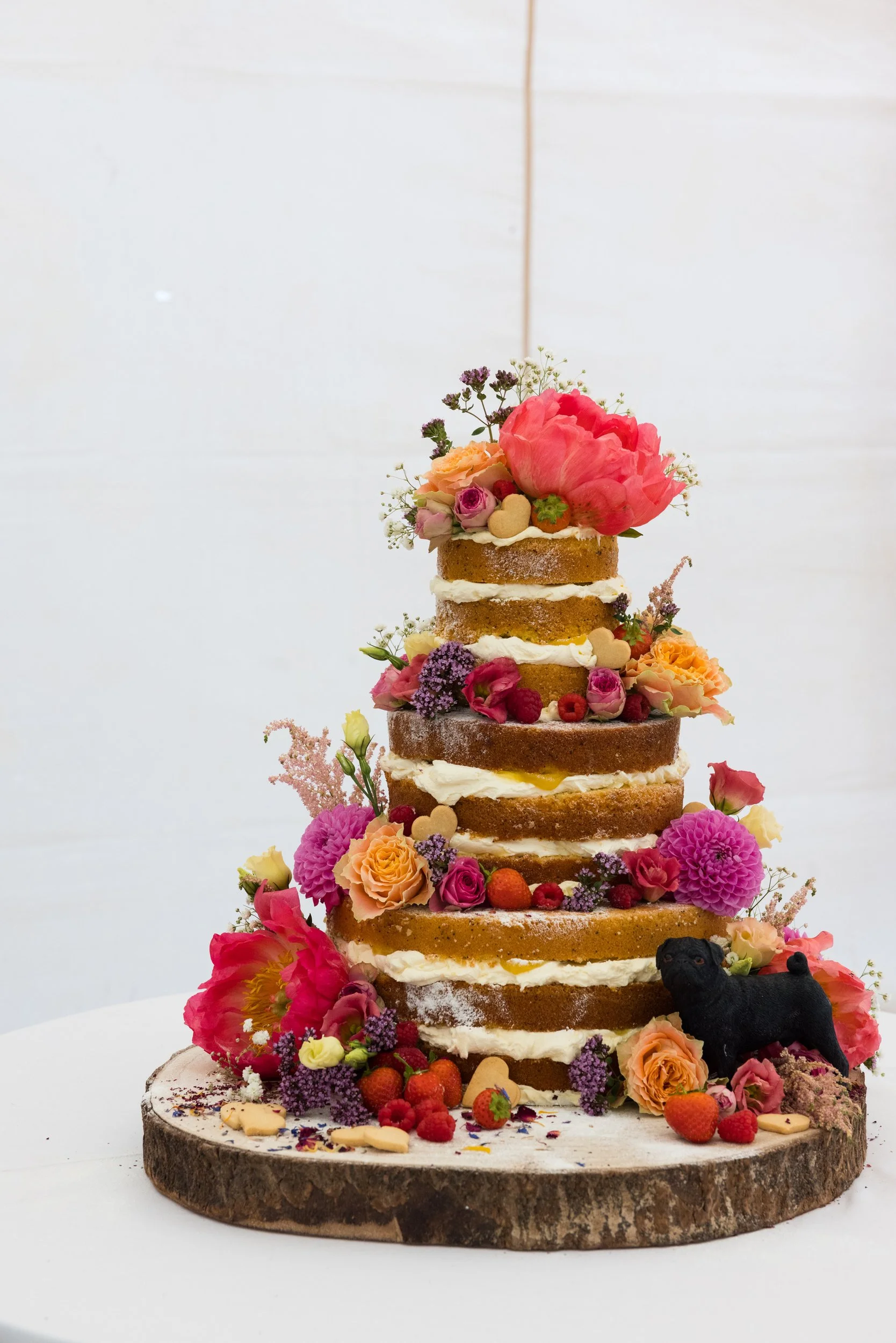 Multi-layered naked cake decorated with fresh flowers, berries, and small animal-shaped cookies, placed on a wooden slab with additional flowers and berries around it.