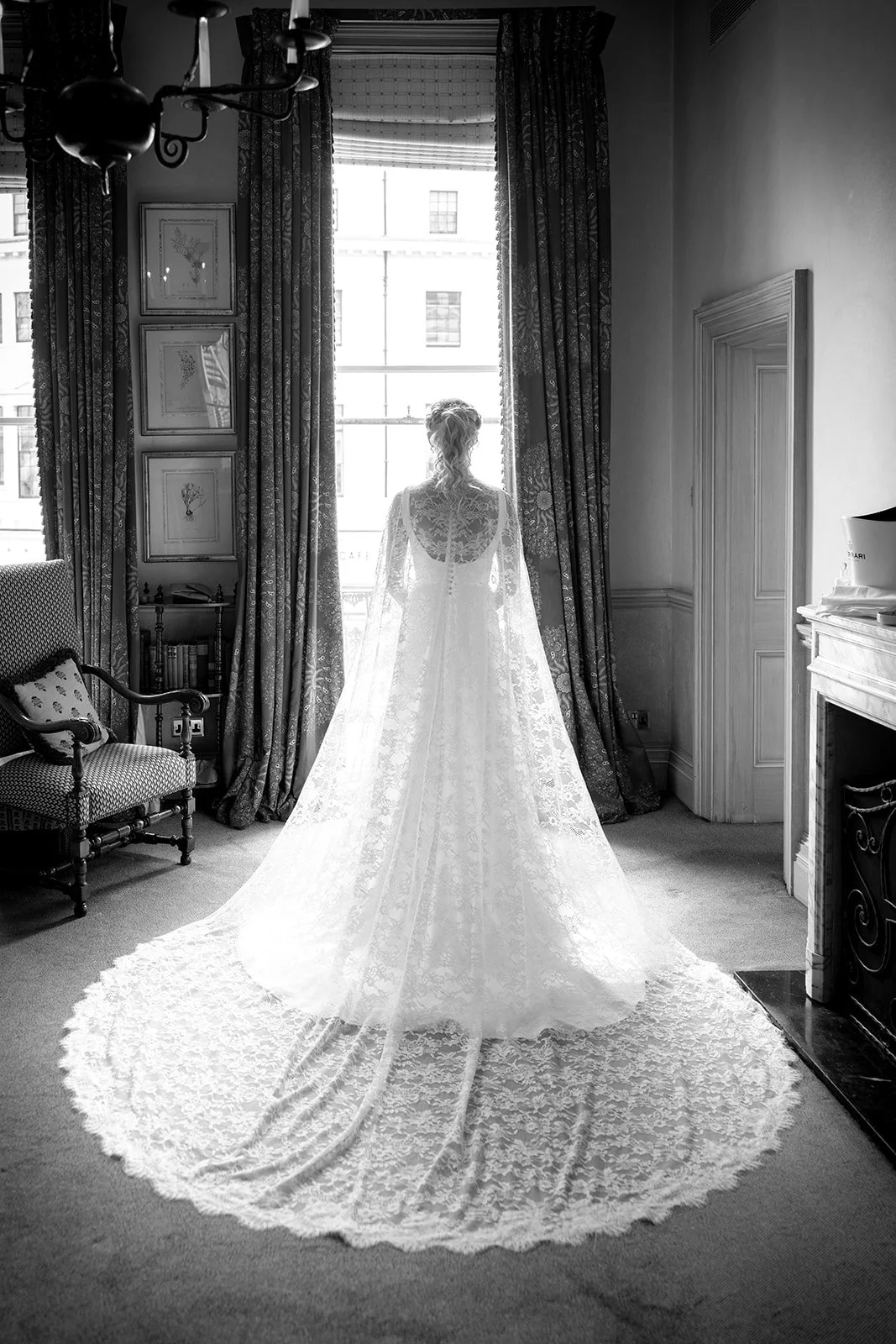 A woman in a wedding dress standing in a room, looking out of a large window with curtains, in black and white.
