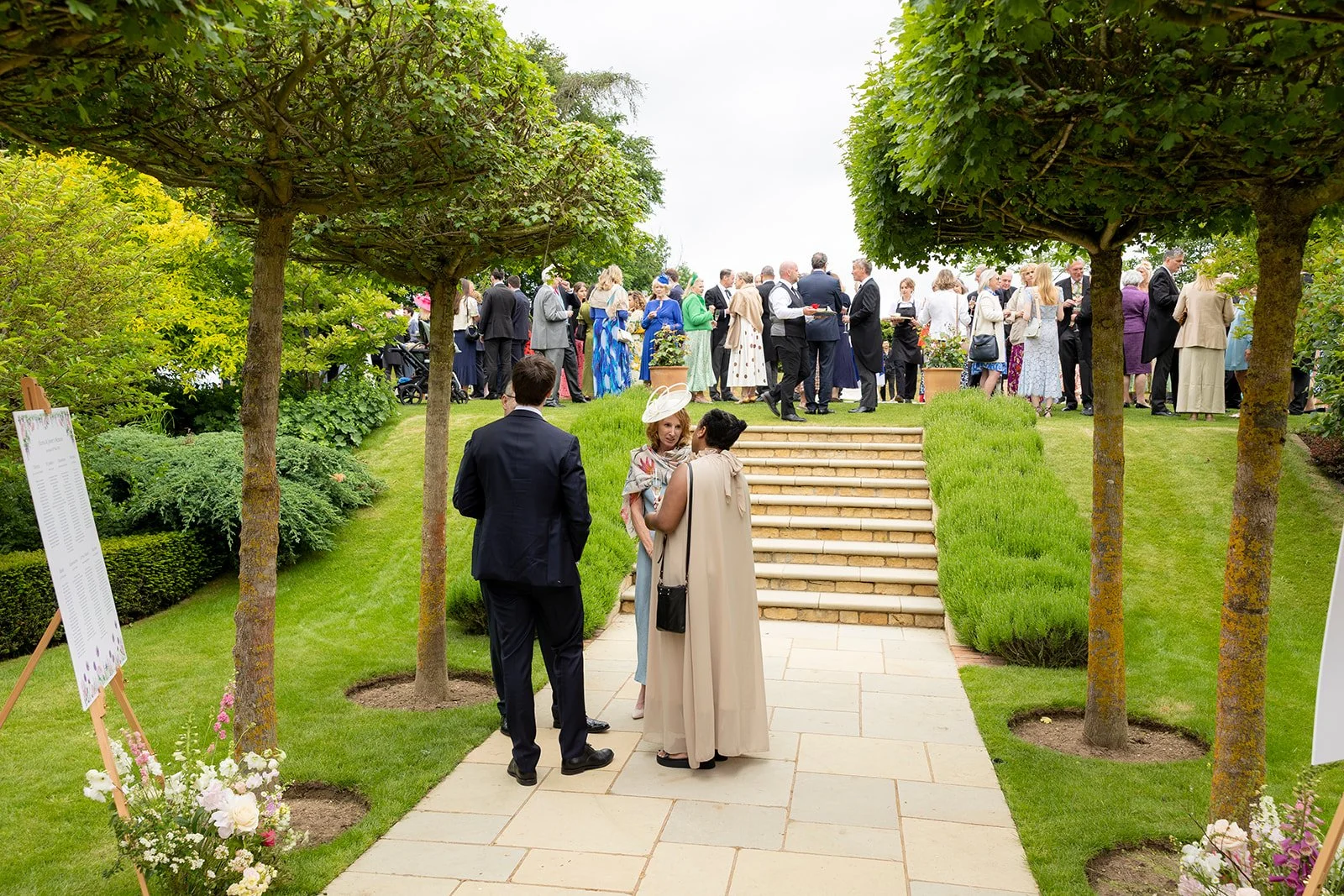Guests gathered outdoors for a garden party or wedding reception, standing and chatting on a lawn with stairs, trees, and flowers.