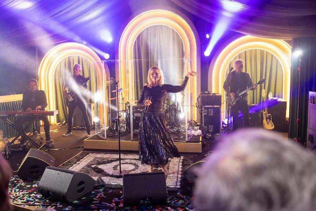 A band performs on stage with a female singer in a black sequined dress holding a microphone. There are keyboard, guitar, and drum instruments, with colorful neon arch-shaped lights in the background and stage lighting creating a vibrant atmosphere.